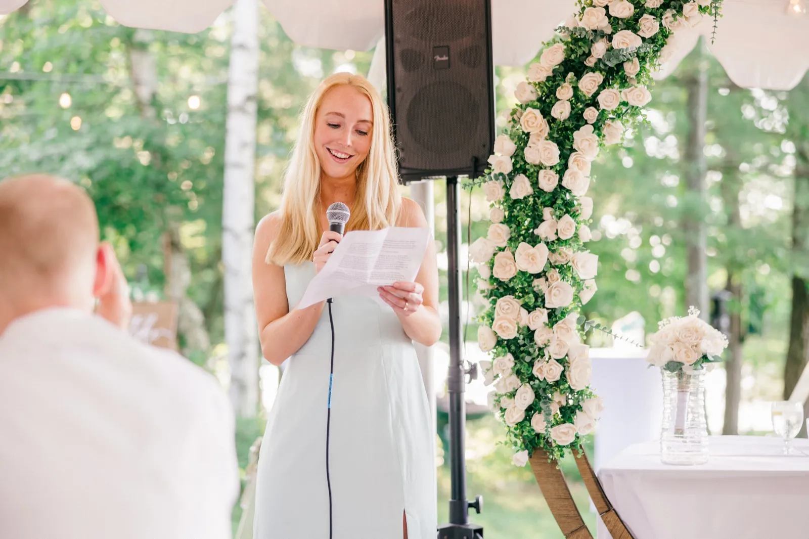 A bridesmaid in a pale blue dress smiles as she reads a speech into a microphone beside a tall floral arch under a reception tent. — Tim Larsen Photography, Brainerd Lakes MN