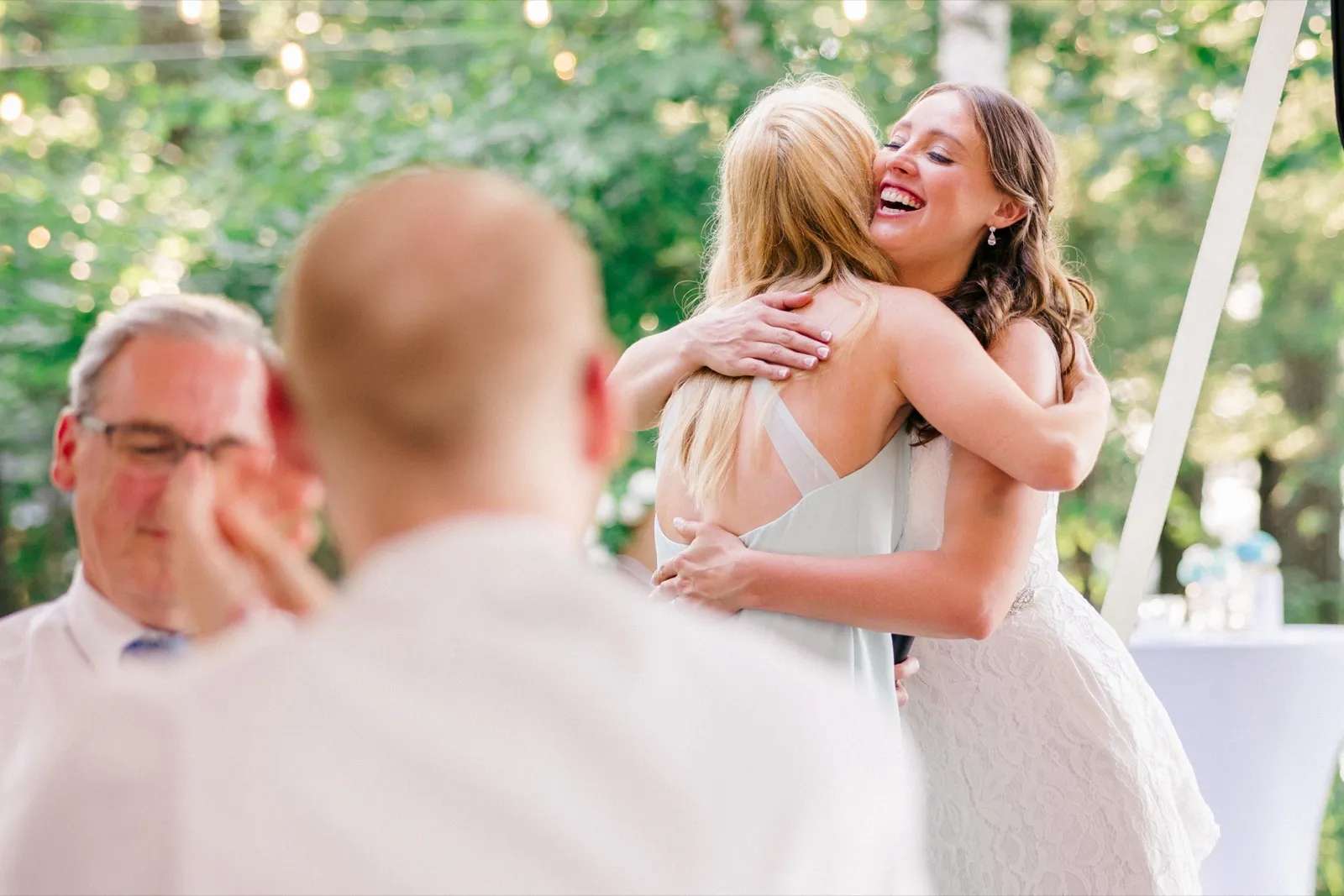 The bride laughs joyfully while embracing a guest, with her father visible out of focus in the foreground wiping a tear. — Tim Larsen Photography, Brainerd Lakes MN