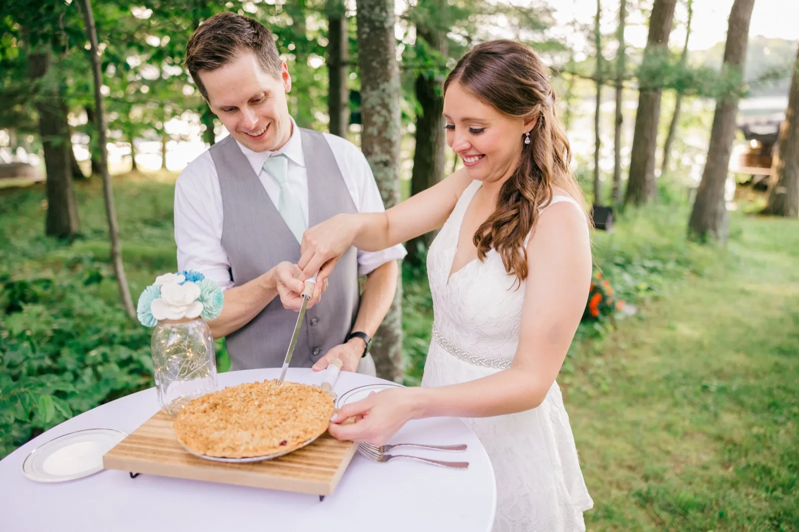 The bride and groom smile while cutting into a crumble-topped pie on a wooden board at an outdoor lakeside reception. — Tim Larsen Photography, Brainerd Lakes MN