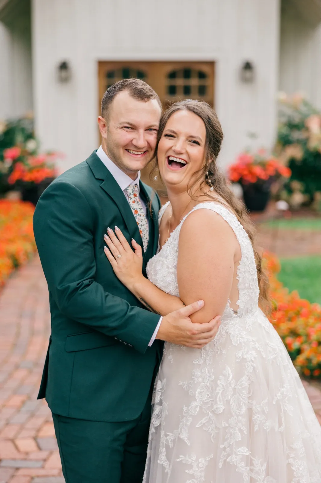 Chelsea and Eric laugh together in an embrace on a brick walkway in front of Grand View Lodge's white facade and flowering garden beds — Tim Larsen Photography, Brainerd Lakes MN