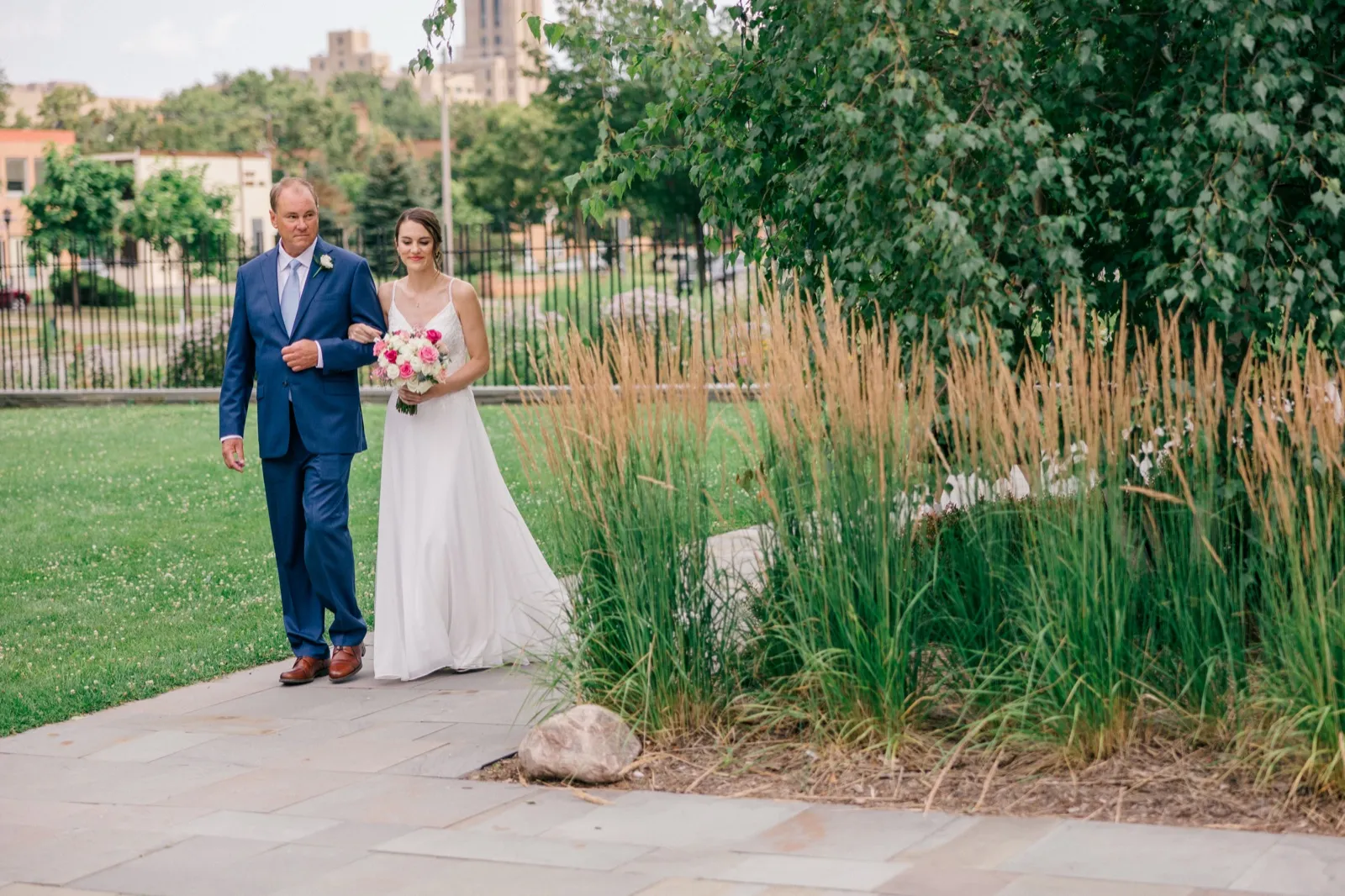 The bride walks arm-in-arm with her father down a stone pathway lined with tall ornamental grasses, the Minneapolis skyline visible in the distance. — Tim Larsen Photography, Minneapolis MN