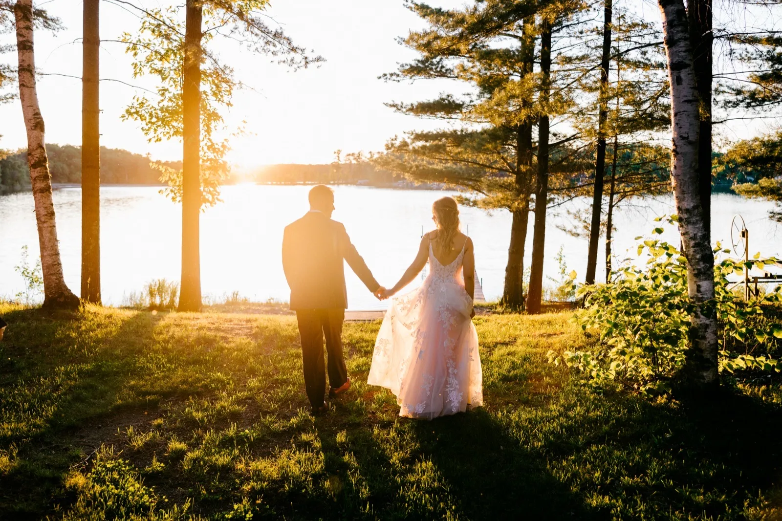 The bride and groom walk hand in hand toward the lake at sunset, framed by tall birch and pine trees with the sun flaring behind them — Tim Larsen Photography, Brainerd Lakes MN