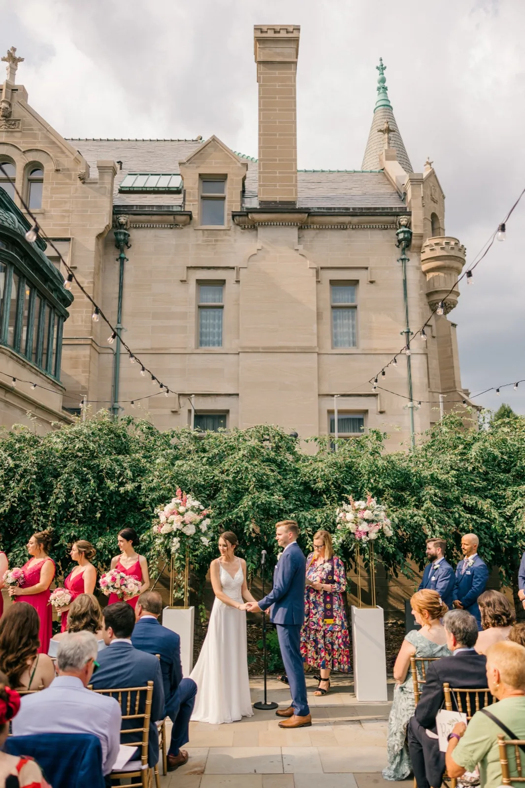 The bride and groom hold hands during their outdoor ceremony in front of the historic American Swedish Institute mansion, surrounded by seated guests and bridal party in pink dresses. — Tim Larsen Photography, Minneapolis MN