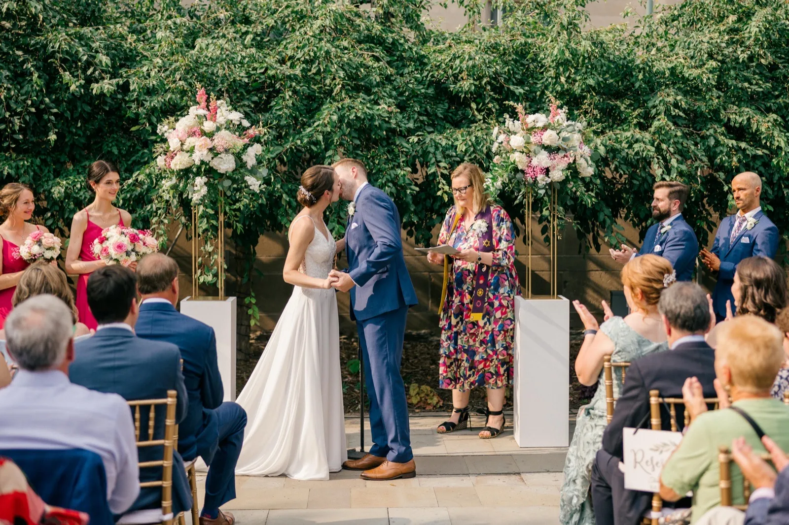 The bride and groom share their first kiss between two tall floral arrangements as the officiant smiles beside them and guests applaud from chairs in the foreground. — Tim Larsen Photography, Minneapolis MN