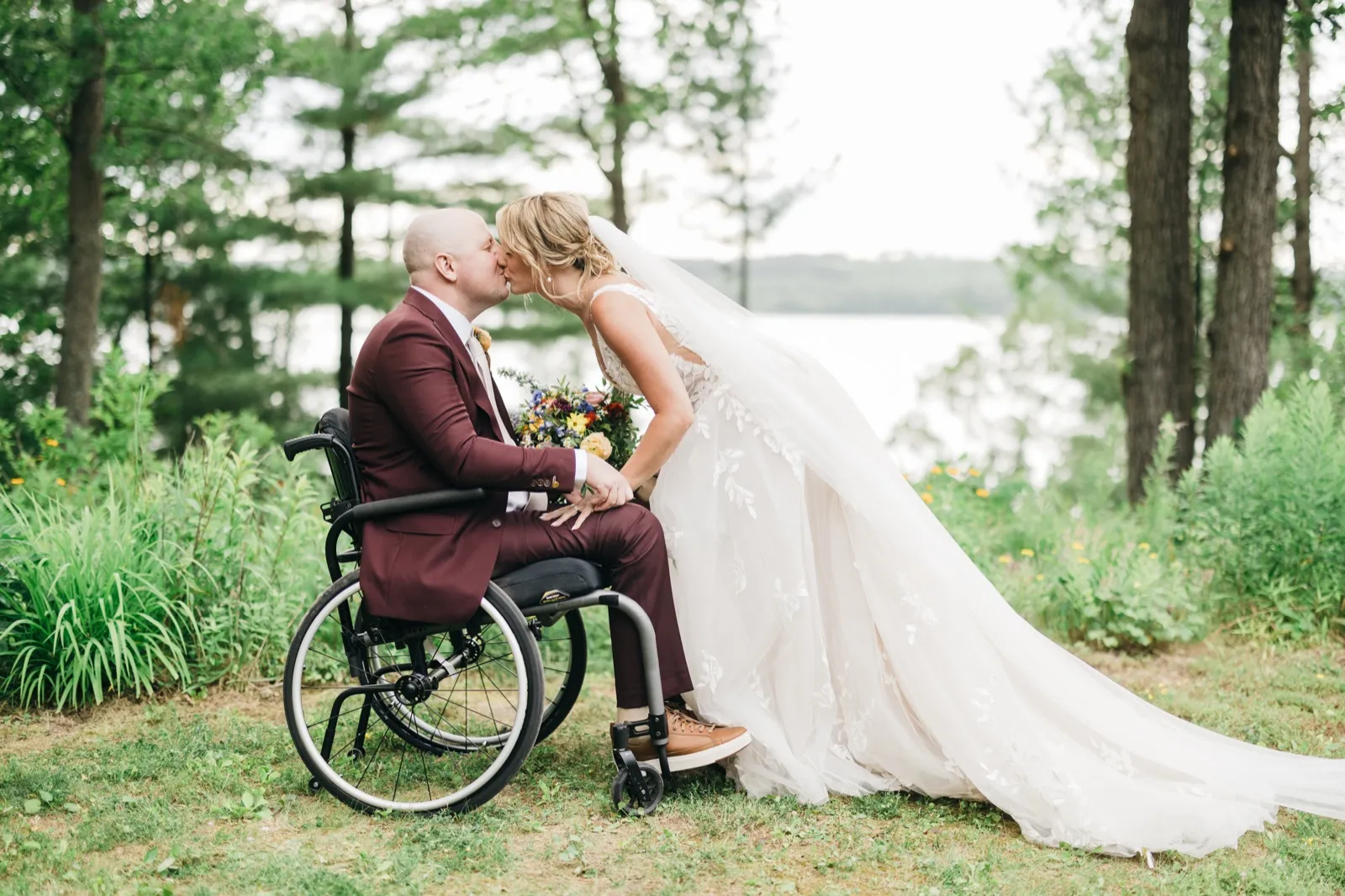 The bride leans down from standing to kiss the groom, who is seated in a wheelchair, while her veil and train sweep across the grass beside a lake lined with pine trees. — Tim Larsen Photography, Brainerd Lakes MN