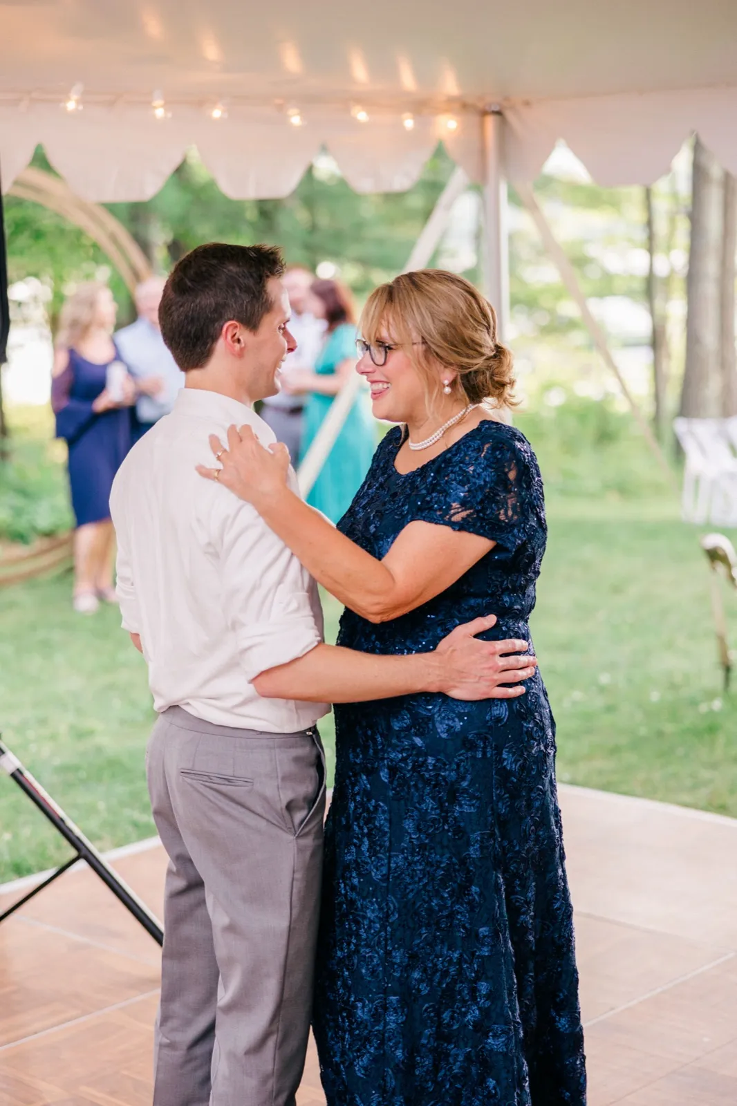The groom's mother smiles up at him as they dance together under a tent strung with cafe lights at a private cabin reception. — Tim Larsen Photography, Brainerd Lakes MN