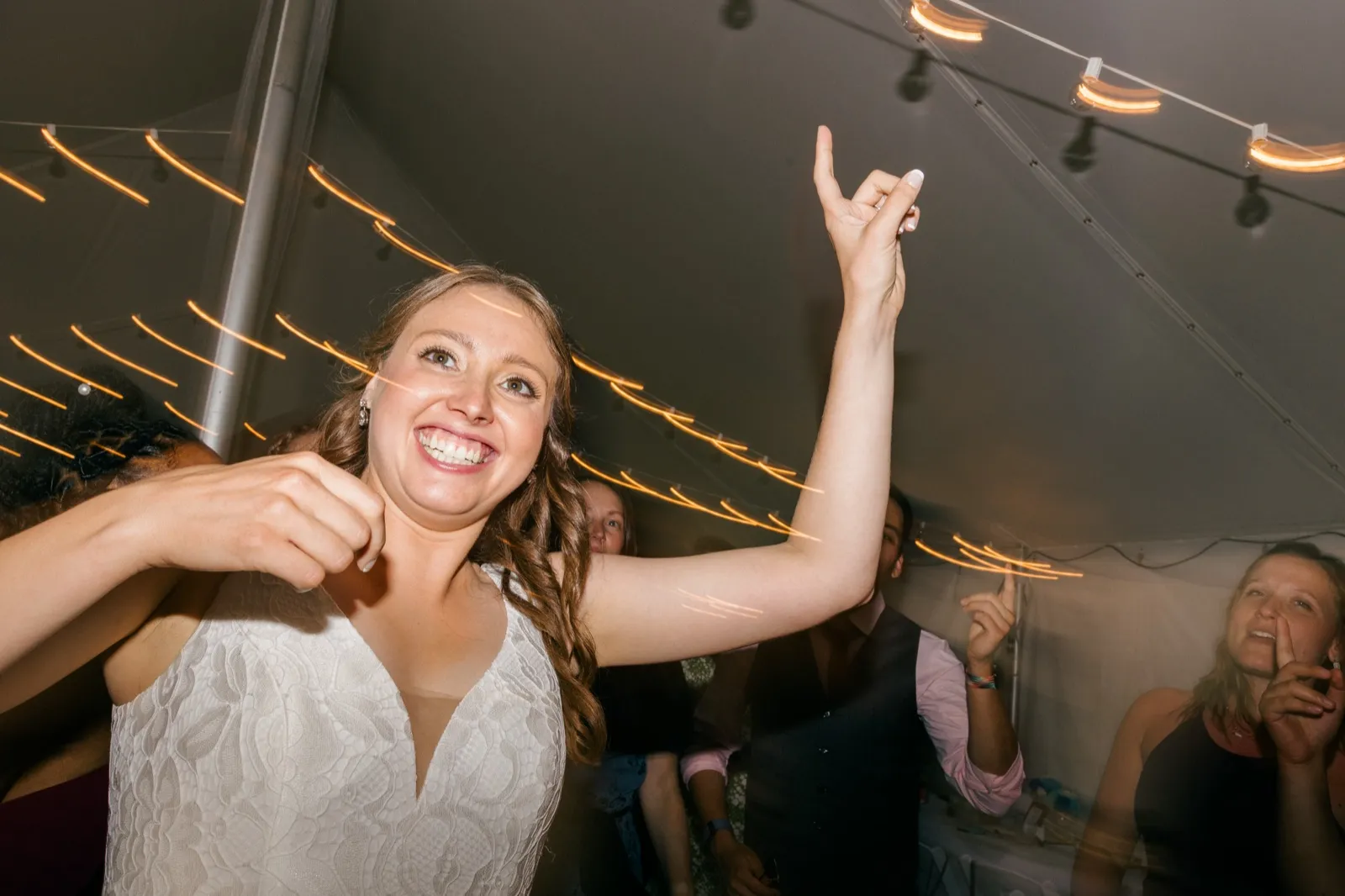 The bride laughs with her arm raised in a rock-on gesture under string lights inside a reception tent, with light streaks trailing through the frame from a dragged shutter. — Tim Larsen Photography, Brainerd Lakes MN