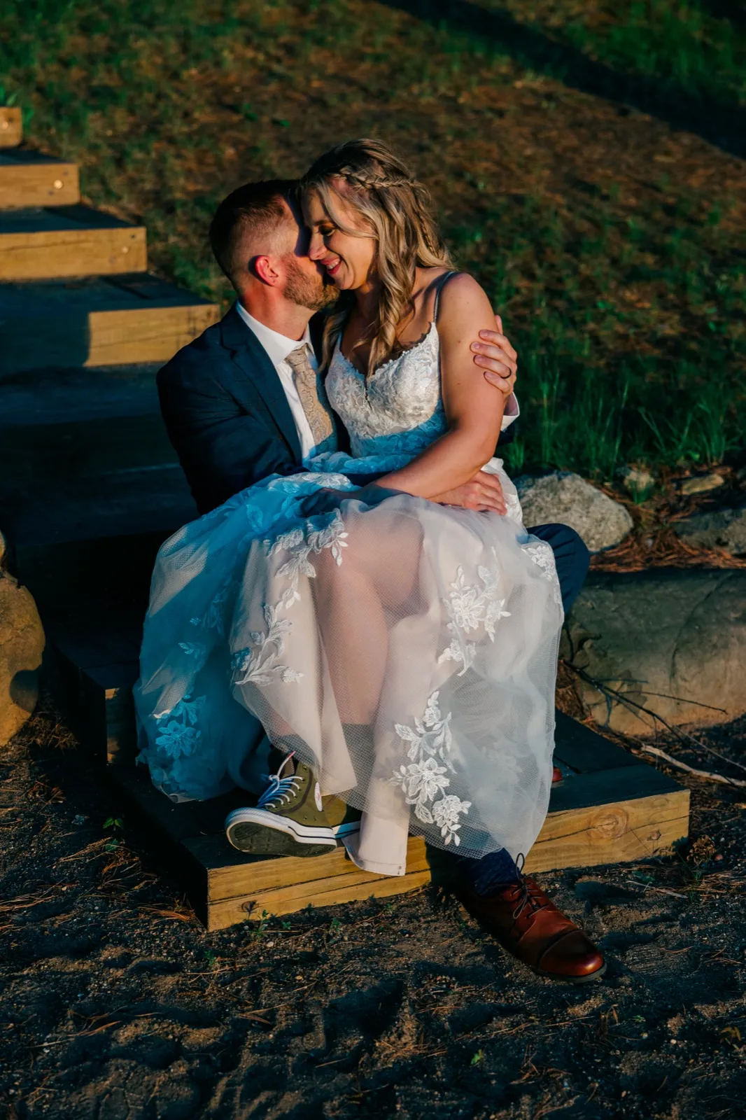 The bride and groom share a quiet kiss while seated on wooden steps at golden hour, her lace dress spilling over the edge and his green sneakers visible beneath — Tim Larsen Photography, Brainerd Lakes MN