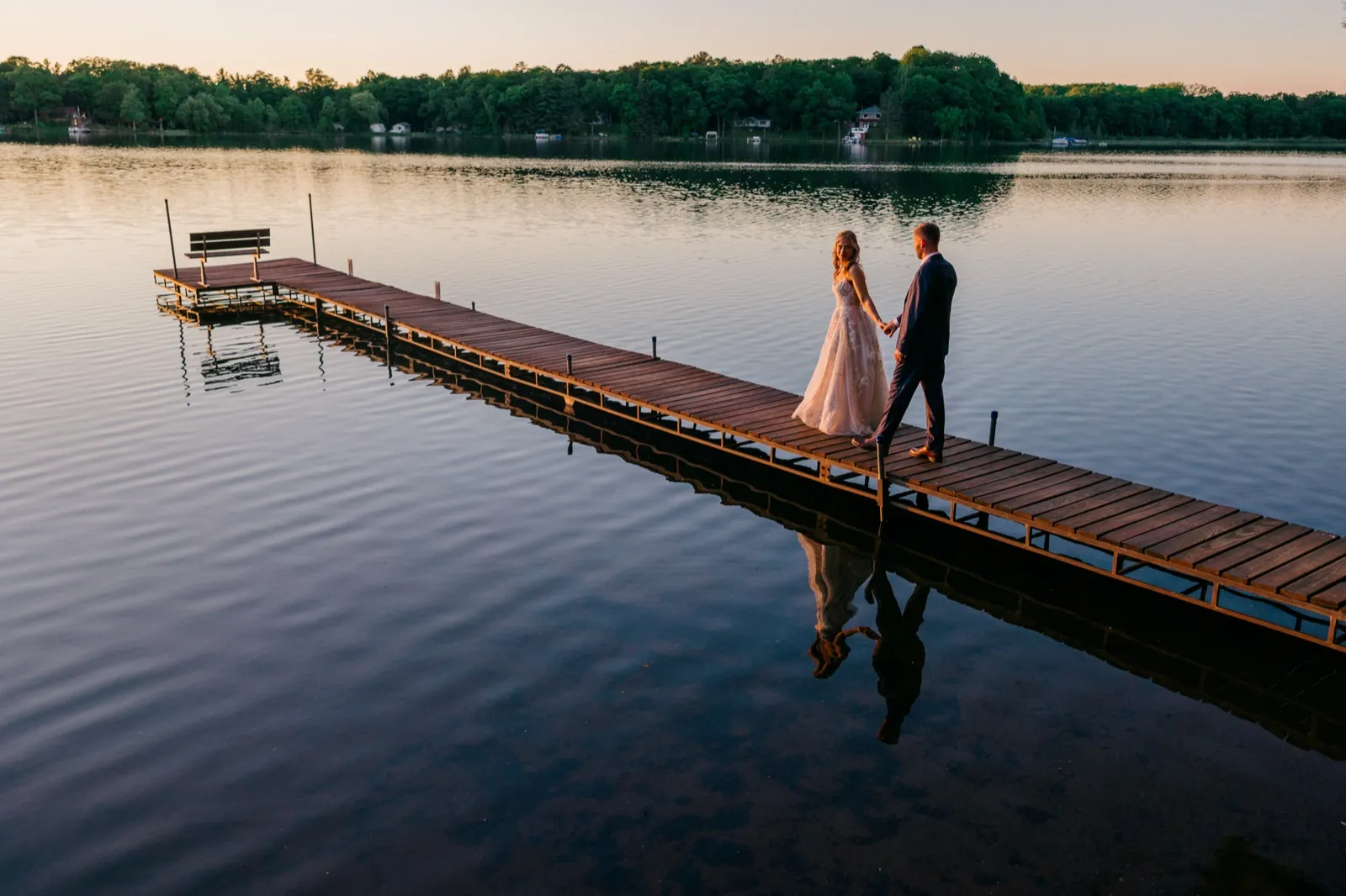 An aerial view of the bride and groom holding hands as they walk along a long wooden dock at dusk, their reflection mirrored in the still lake below — Tim Larsen Photography, Brainerd Lakes MN