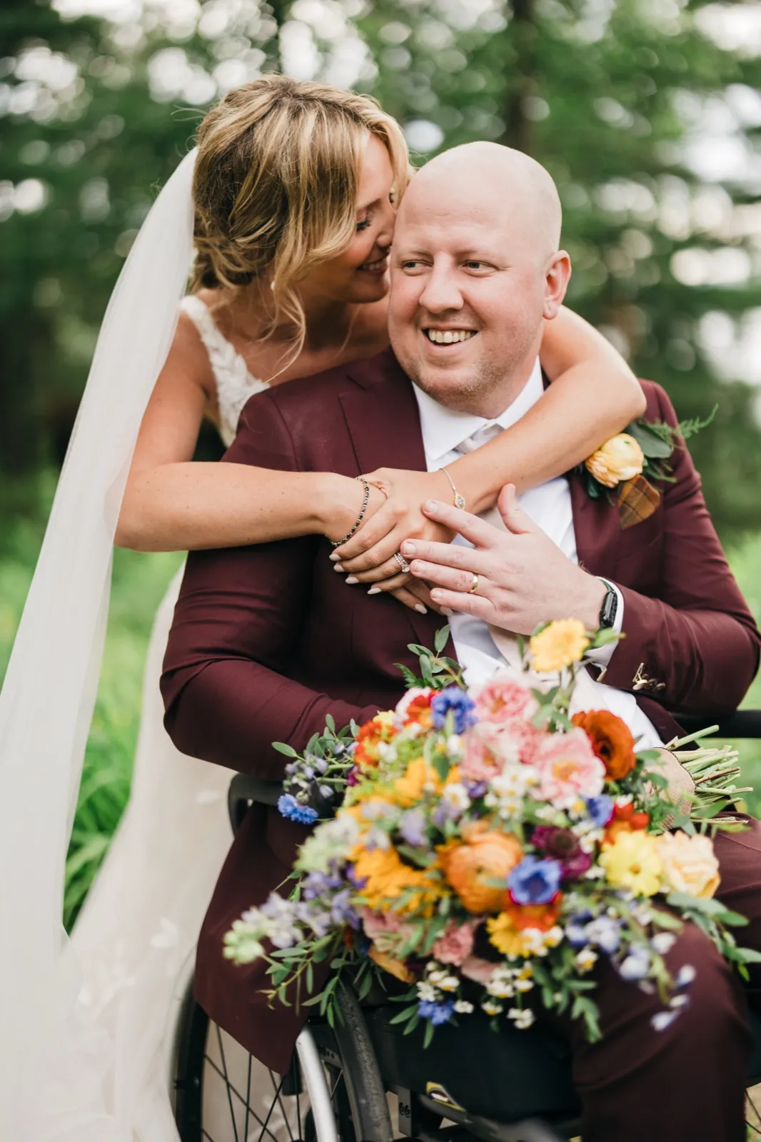 The bride leans over her groom, who is seated in a wheelchair, pressing her face to his cheek as he smiles broadly while holding a vivid wildflower bouquet. — Tim Larsen Photography, Brainerd Lakes MN