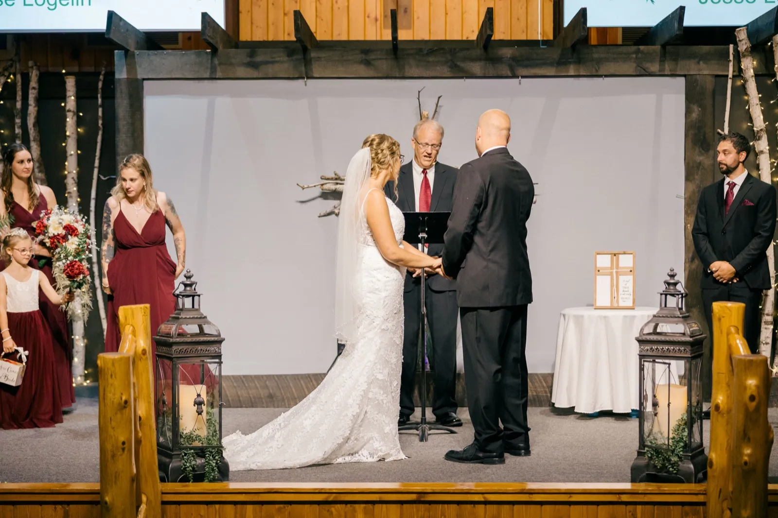 Kim and Jesse hold hands facing the officiant on a wooden ceremony stage flanked by birch trunks and large lanterns, with bridesmaids and groomsmen watching — Tim Larsen Photography, Brainerd Lakes MN