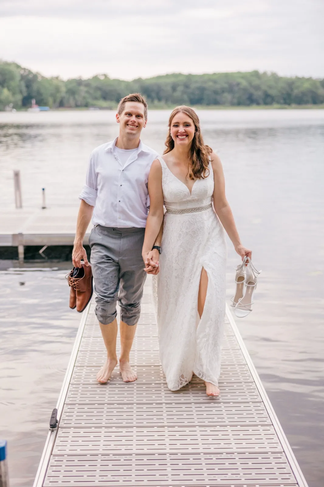 The bride and groom walk barefoot down a dock holding their shoes, smiling at the camera with an overcast lake behind them. — Tim Larsen Photography, Brainerd Lakes MN