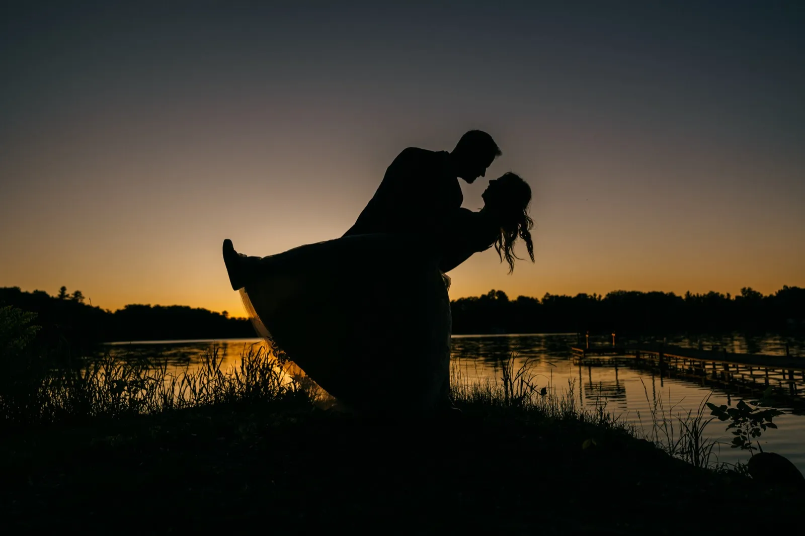 The groom dips the bride into a near-kiss as their silhouettes stand against a glowing dusk sky over a Brainerd Lakes lakeshore with a dock extending into the water — Tim Larsen Photography, Brainerd Lakes MN