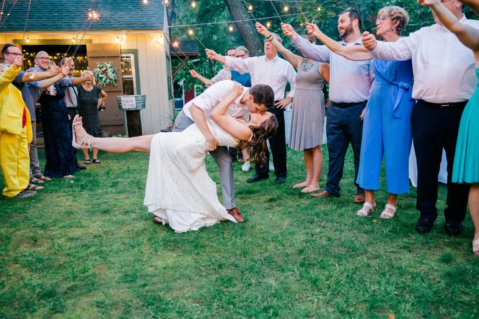 The bride is dipped backward in a kiss with the groom as wedding guests hold sparklers overhead in a backyard at dusk. — Tim Larsen Photography, Brainerd Lakes MN