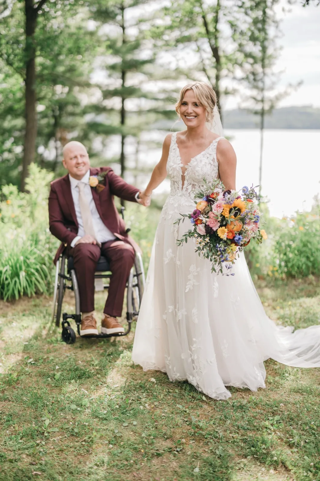 The bride beams while holding a vibrant wildflower bouquet and the groom's hand as he sits in his wheelchair behind her on a wooded lakeside lawn. — Tim Larsen Photography, Brainerd Lakes MN