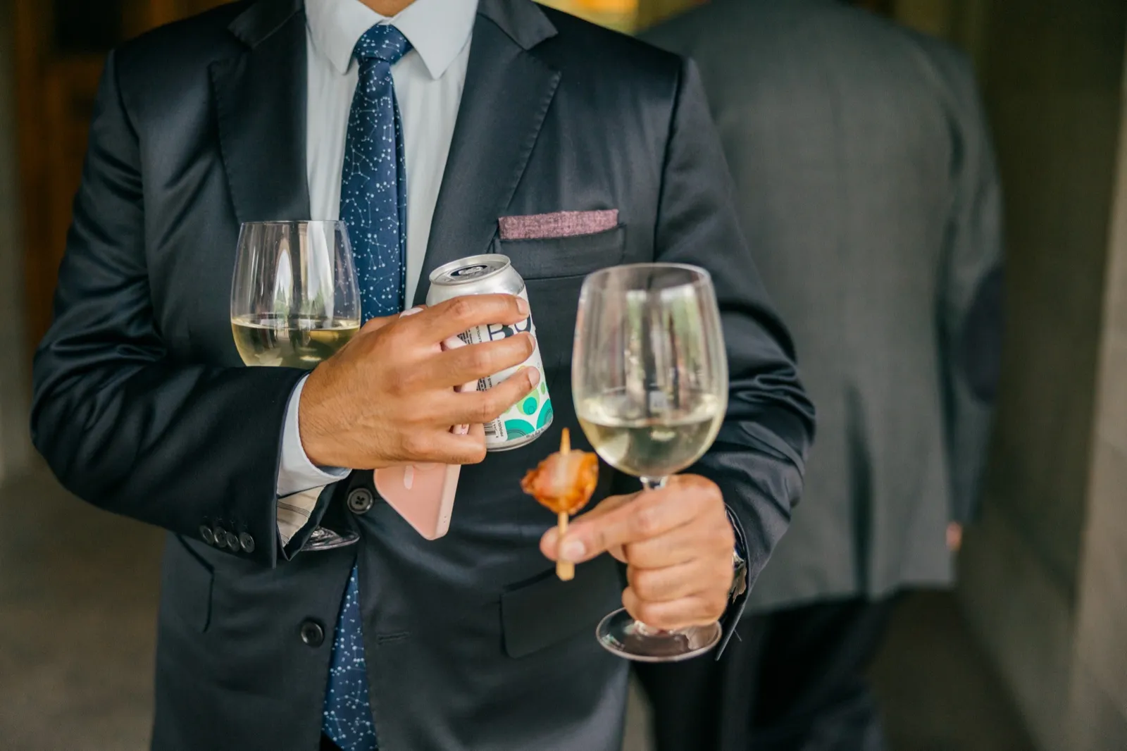 A guest in a navy suit and constellation-patterned tie holds a glass of white wine, a seltzer can, a phone, and a skewered appetizer all at once. — Tim Larsen Photography, Minneapolis MN
