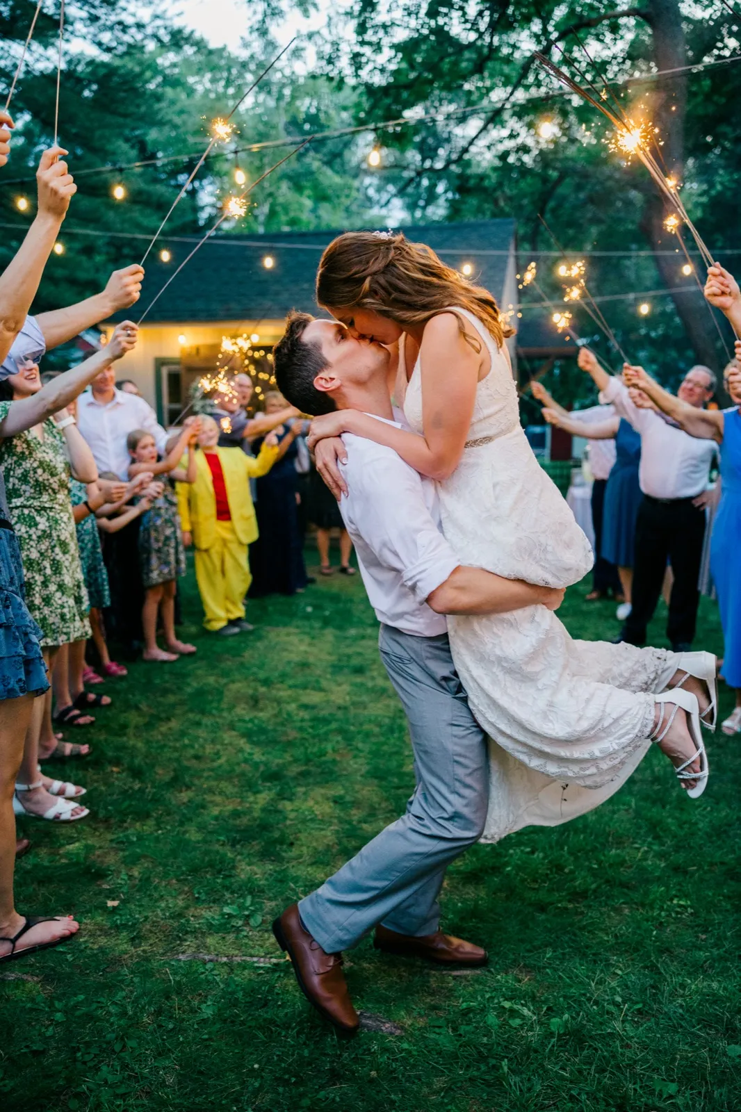 The groom lifts the bride off the ground for a kiss inside a tunnel of guests holding sparklers in a backyard at dusk. — Tim Larsen Photography, Brainerd Lakes MN