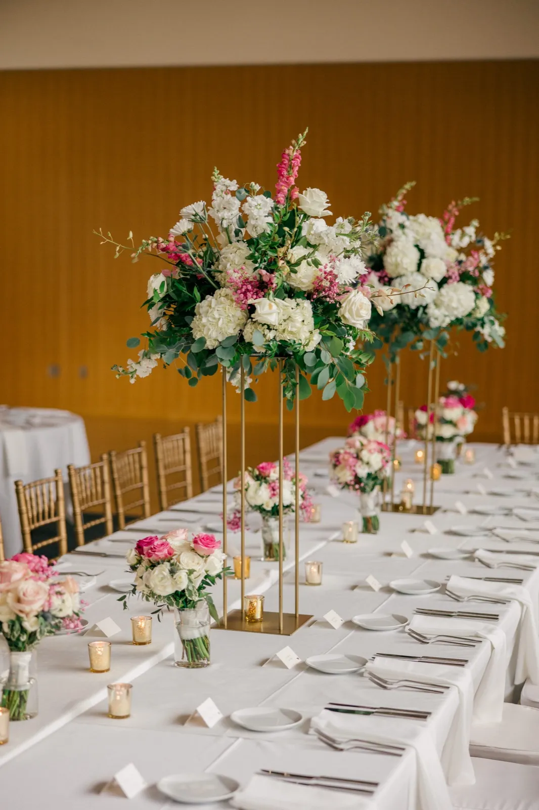 A long reception table at the American Swedish Institute set with white linens, gold chiavari chairs, votive candles, and a tall pink and white floral centerpiece on a gold stand. — Tim Larsen Photography, Minneapolis MN