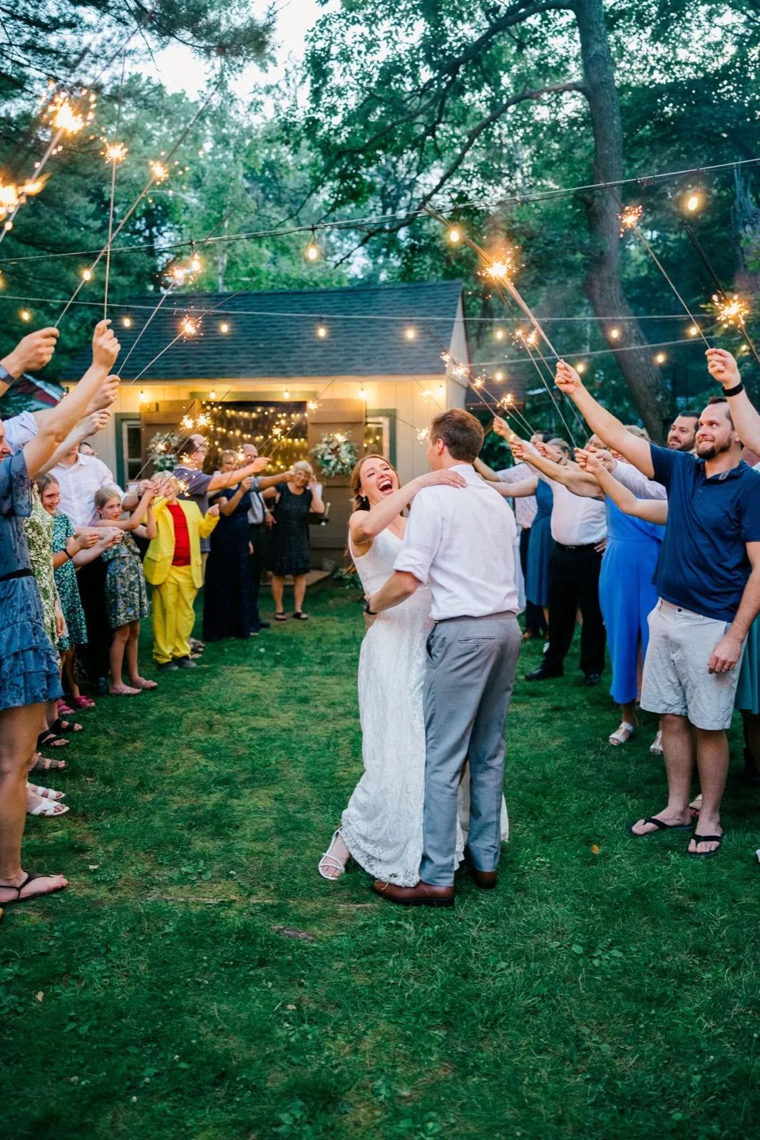 The bride throws her head back laughing in her groom's arms as guests form a sparkler tunnel around them in front of a string-lit cabin at dusk. — Tim Larsen Photography, Brainerd Lakes MN