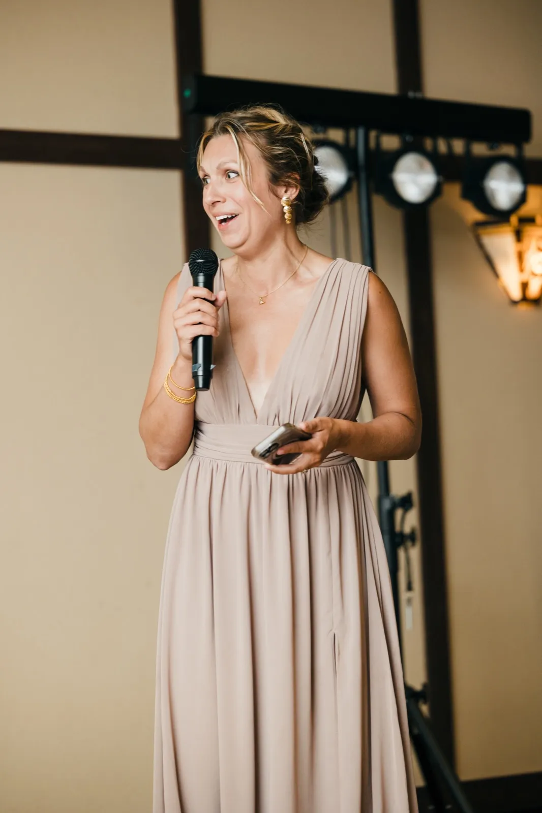 A bridesmaid in a champagne gown holds a microphone and notecards, eyes wide and mouth open mid-anecdote during a reception toast.