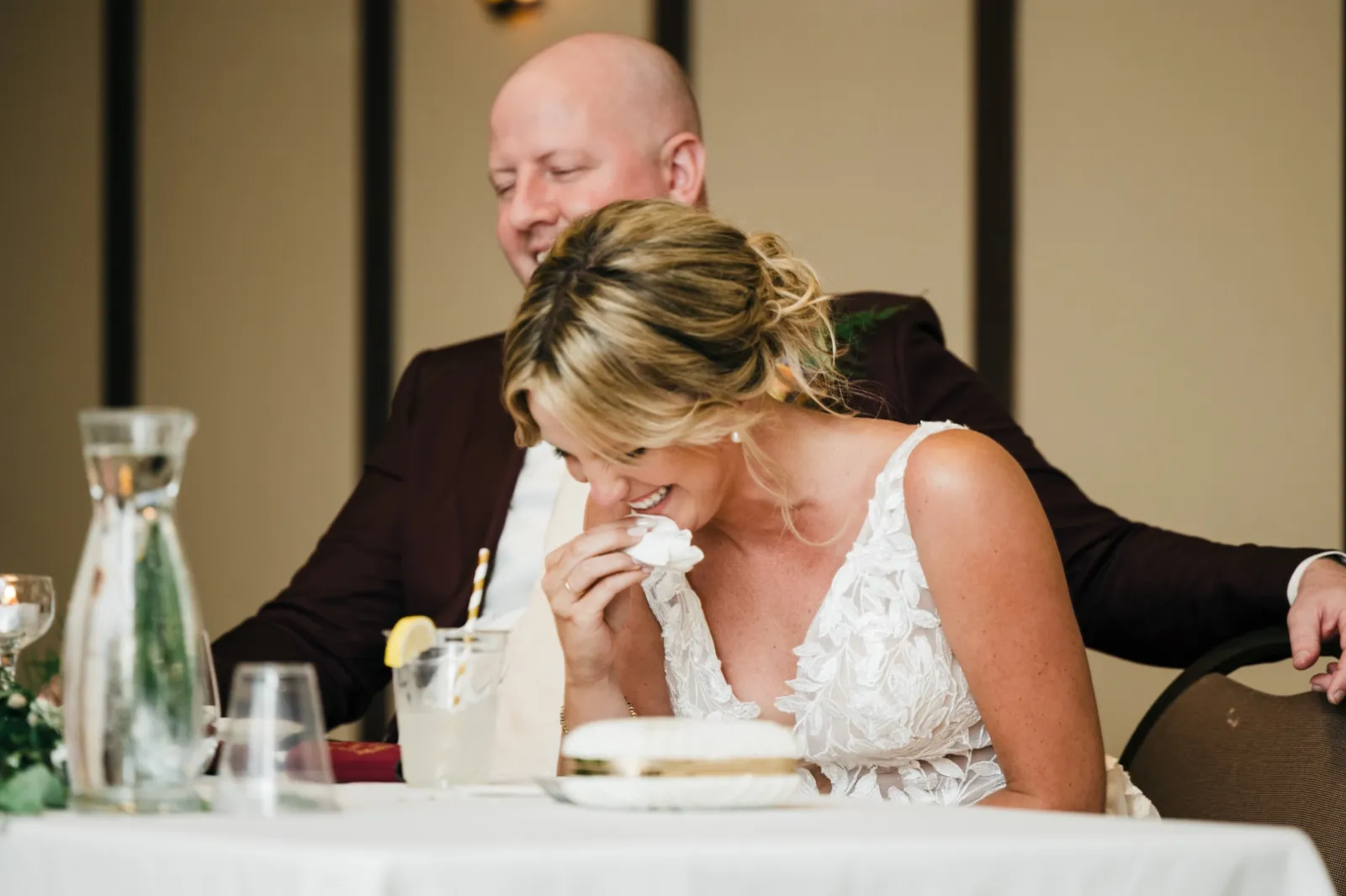 The bride wipes her eyes with a napkin and laughs through tears while the groom watches her with a soft smile from behind at their sweetheart table. — Tim Larsen Photography, Brainerd Lakes MN