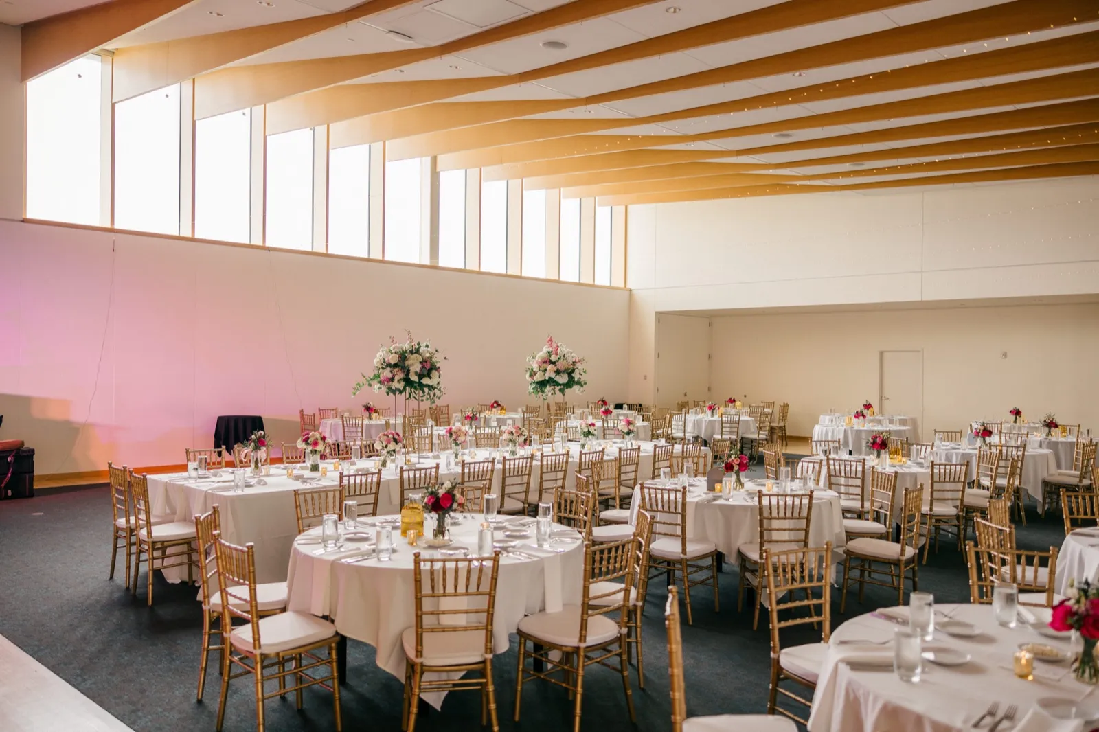 An empty reception hall at the American Swedish Institute set with round tables, gold chiavari chairs, and tall pink and white floral centerpieces beneath a wood-slatted ceiling. — Tim Larsen Photography, Minneapolis MN