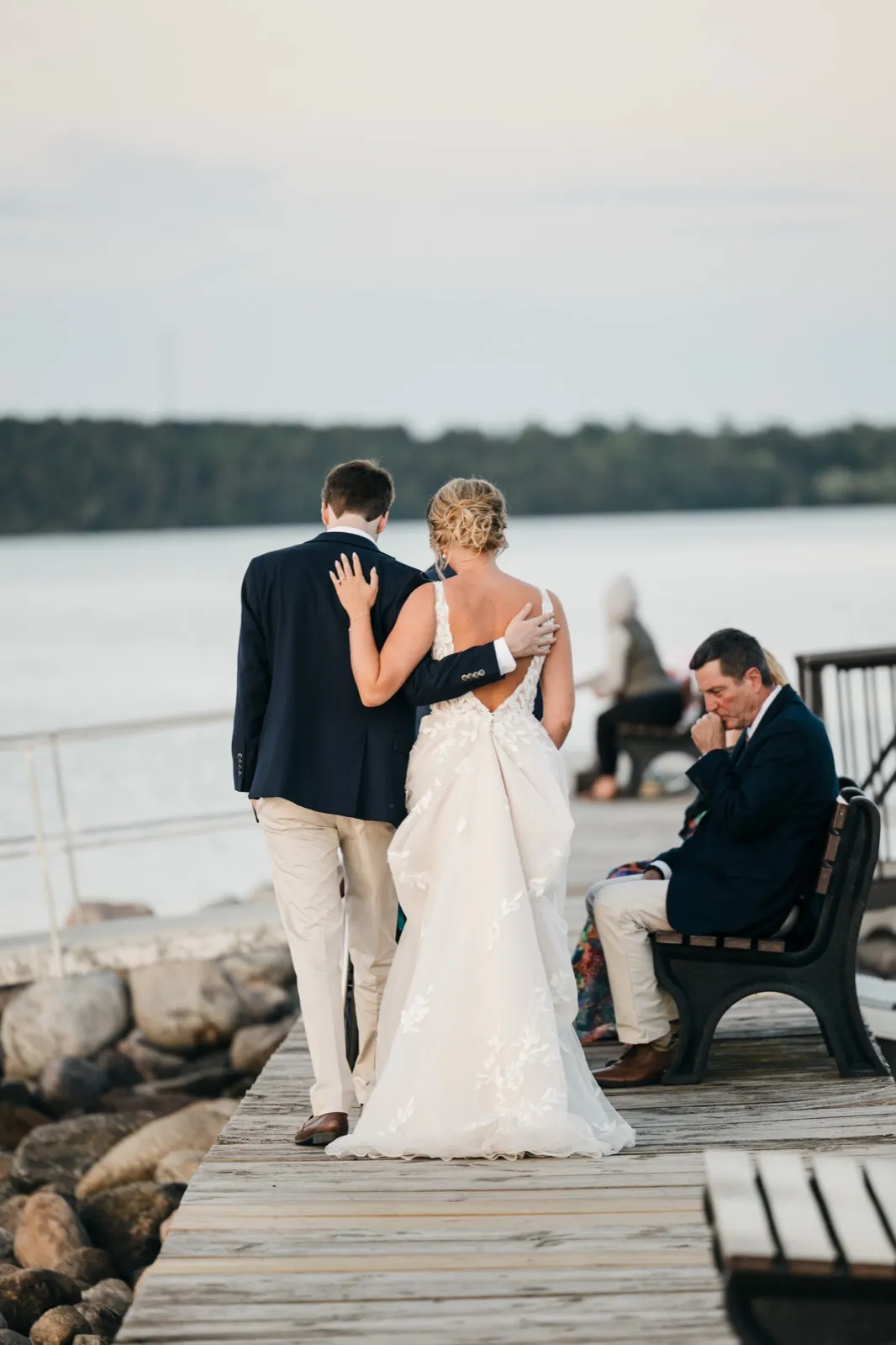 Jenna walks arm-in-arm with her father down the marina dock at dusk, a seated guest in the background quietly wiping his eyes. — Tim Larsen Photography, Brainerd Lakes MN