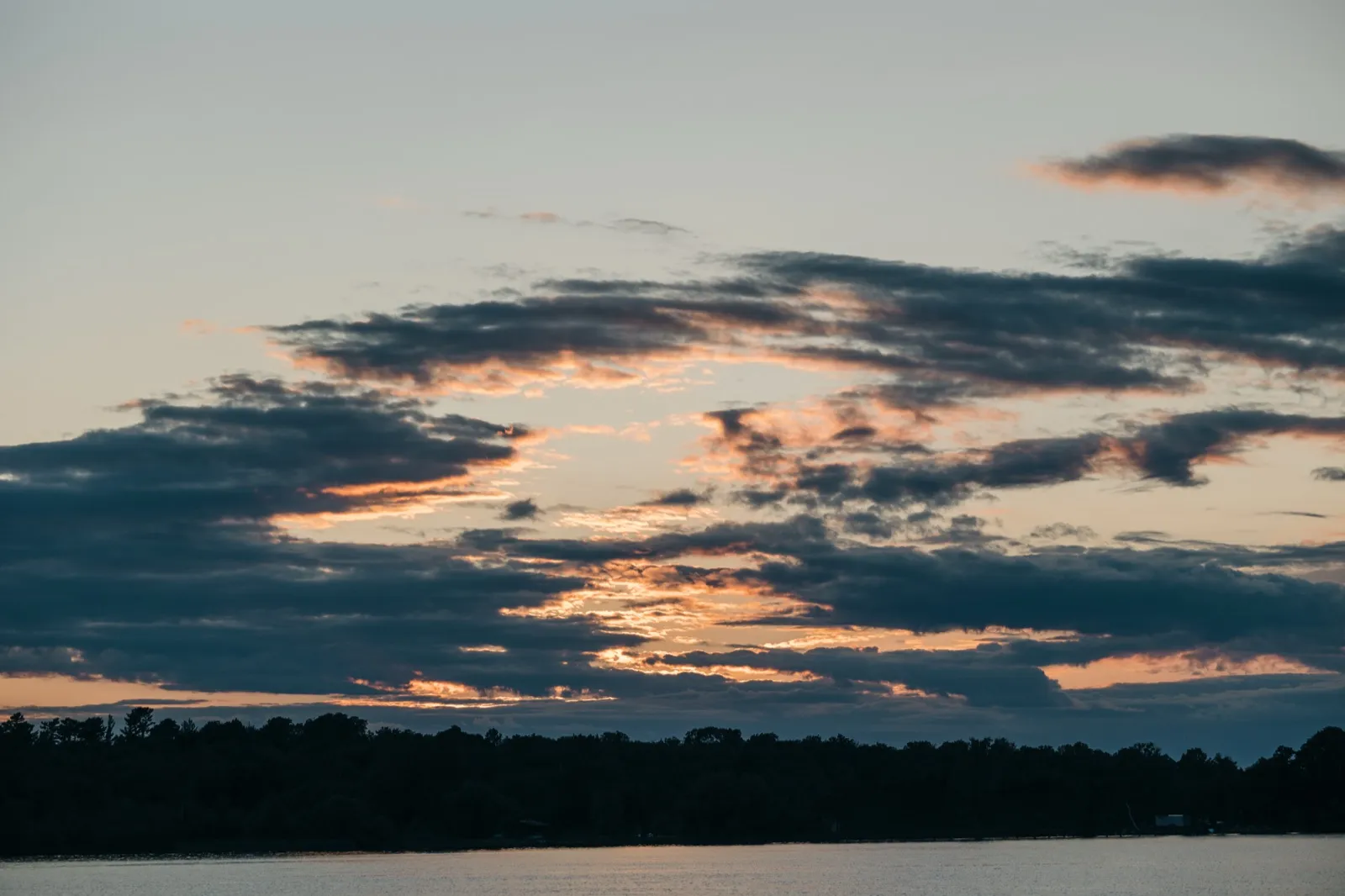 A wide sunset sky streaked with peach and slate clouds stretches above a dark treeline and calm lake water. — Tim Larsen Photography, Brainerd Lakes MN