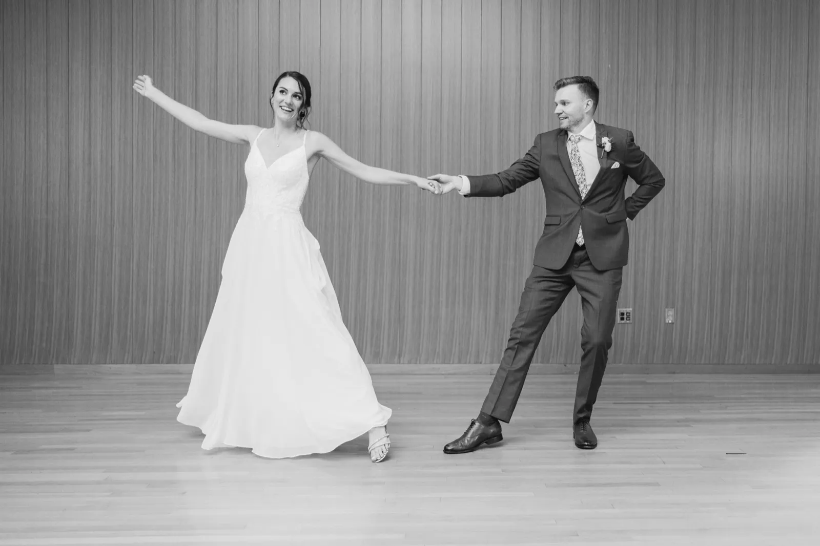 The bride and groom stretch their arms apart while holding hands in a playful dance pose on a wood floor against a paneled wall. — Tim Larsen Photography, Minneapolis MN