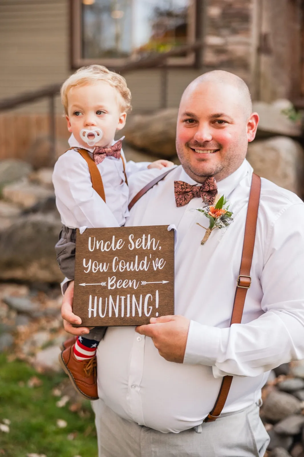 The groom holds his young nephew, who wears a bowtie and holds a pacifier, while the pair display a wooden sign reading 'Uncle Seth, You Could've Been Hunting!' outside the lodge — Tim Larsen Photography, Brainerd Lakes MN