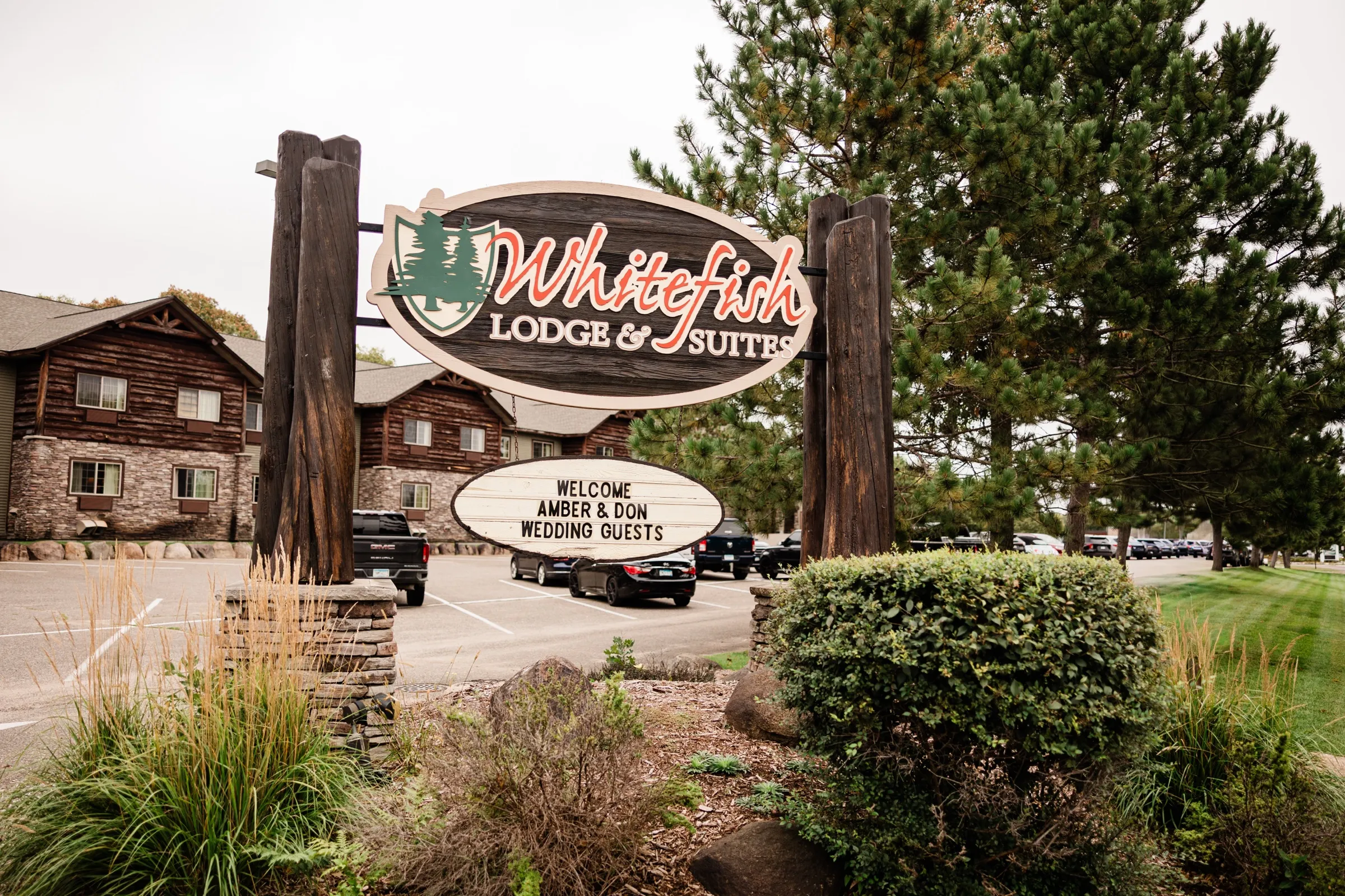 The carved Whitefish Lodge & Suites entrance sign hangs between log posts with a welcome marquee for the wedding guests, log-and-stone lodge buildings and pines behind it. — Tim Larsen Photography, Brainerd Lakes MN