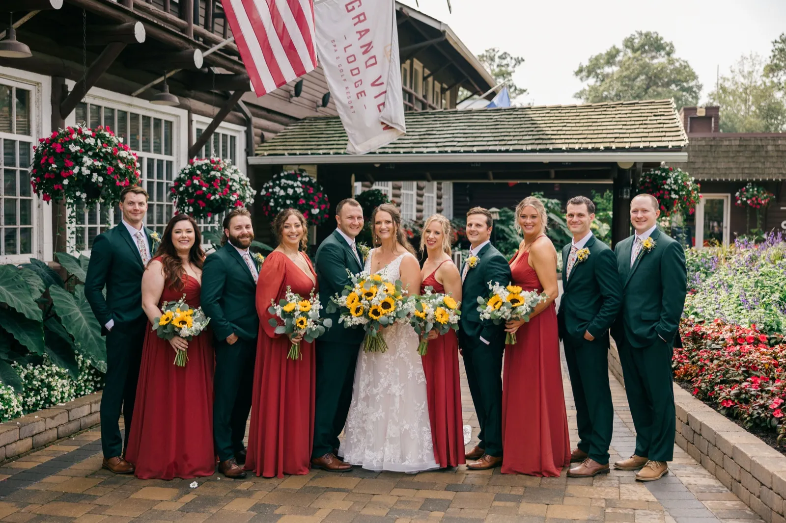Chelsea and Eric stand center with their bridal party in red dresses and dark suits, holding sunflower bouquets in front of the Grand View Lodge entrance — Tim Larsen Photography, Brainerd Lakes MN
