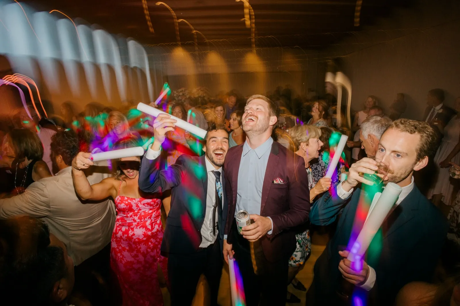 The groom laughs with his head thrown back while guests around him wave colored foam glow sticks on a packed dance floor. — Tim Larsen Photography, Minneapolis MN