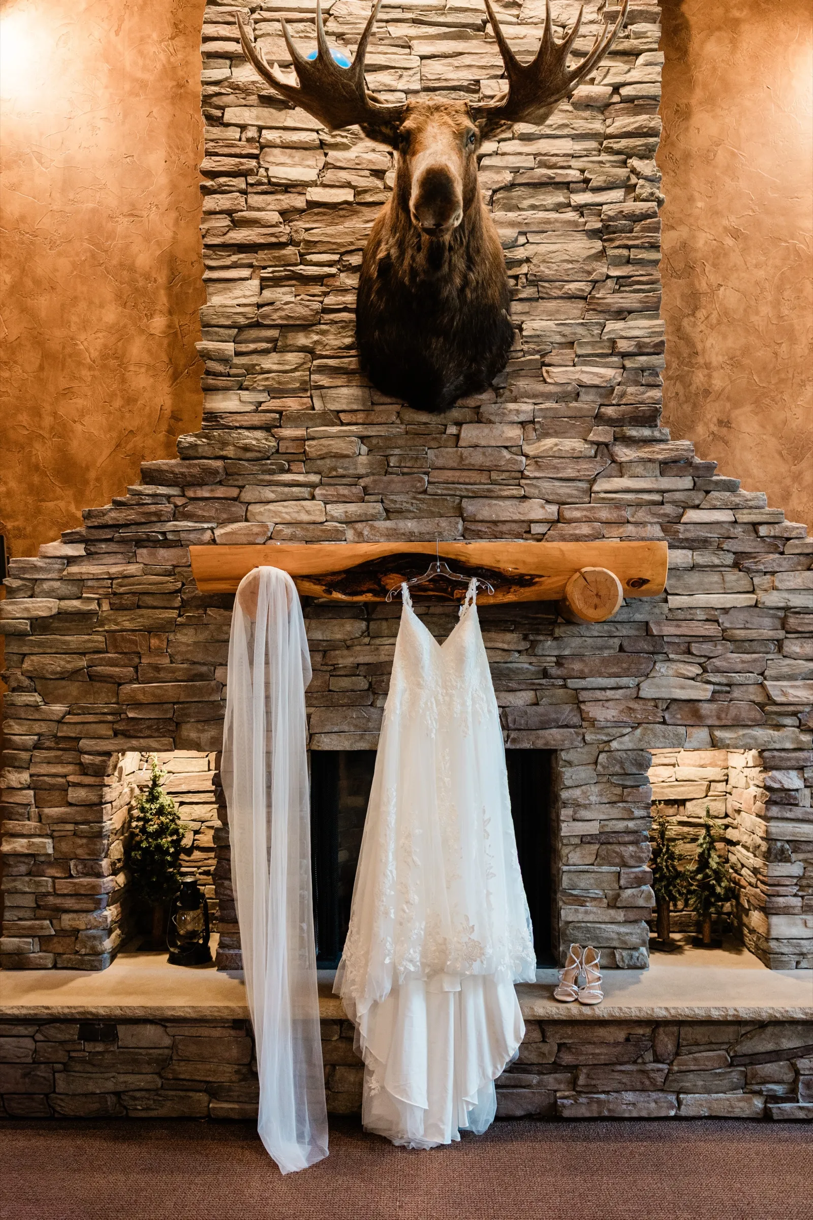 A lace wedding gown and long veil hang from a log mantel on a stacked-stone fireplace under a mounted moose head, with strappy heels on the hearth. — Tim Larsen Photography, Brainerd Lakes MN