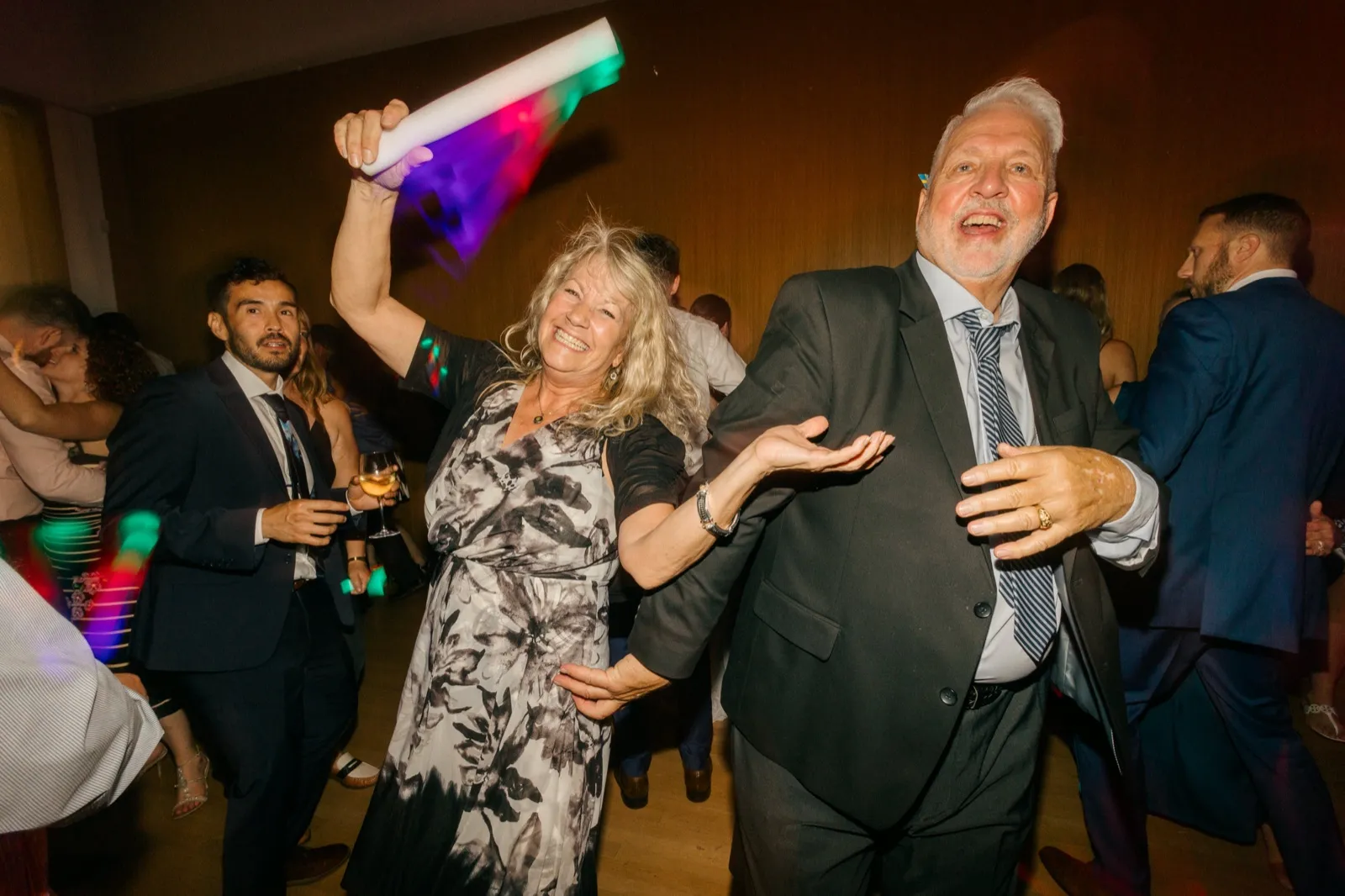 A woman in a floral dress waves a foam glow stick overhead while dancing beside a man in a gray suit on a crowded reception dance floor lit with colored lights. — Tim Larsen Photography, Minneapolis MN