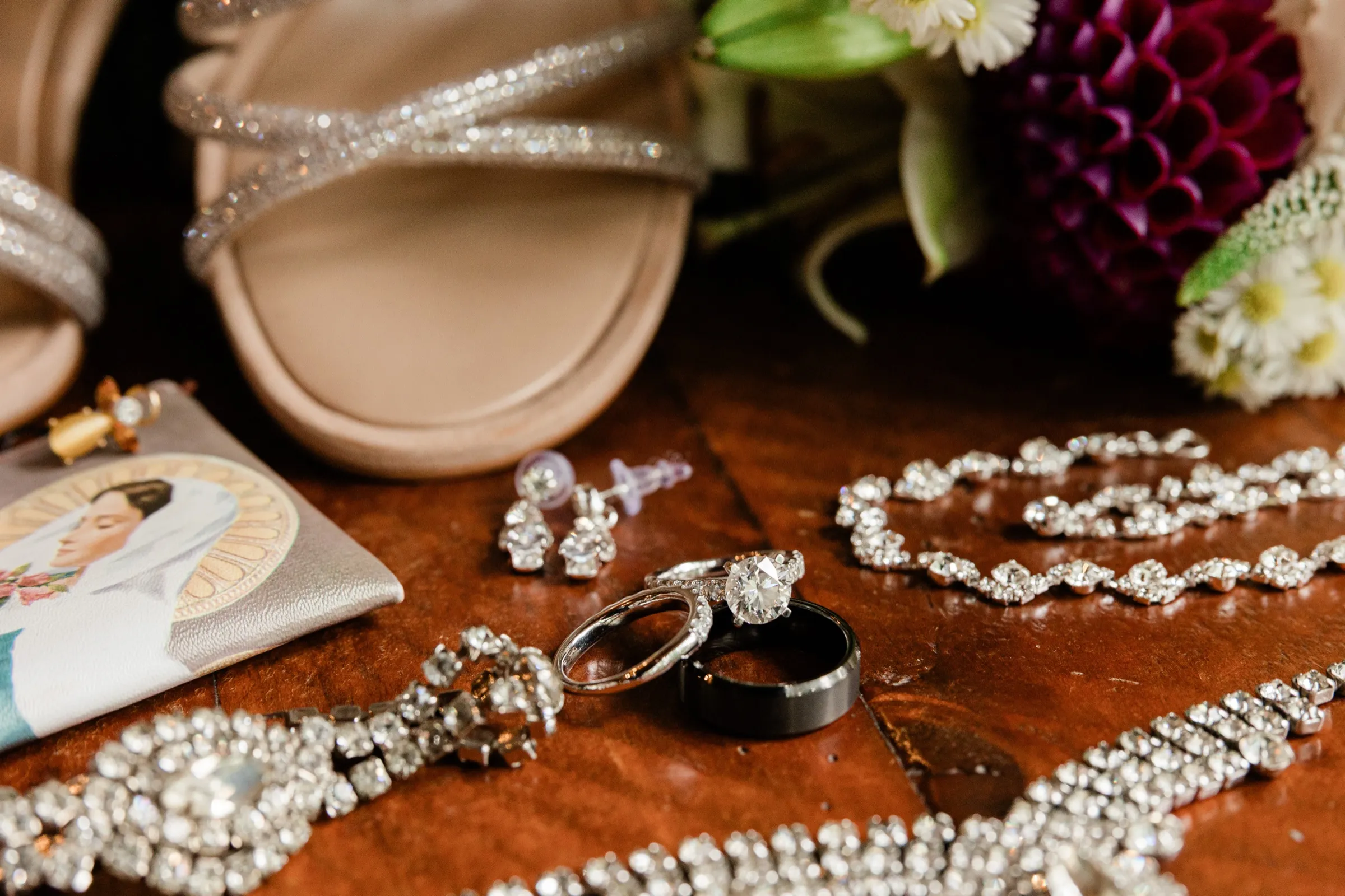 A diamond engagement ring rests on a silver band and a black men's band, surrounded by rhinestone jewelry and the toe of a glittering heel. — Tim Larsen Photography, Brainerd Lakes MN