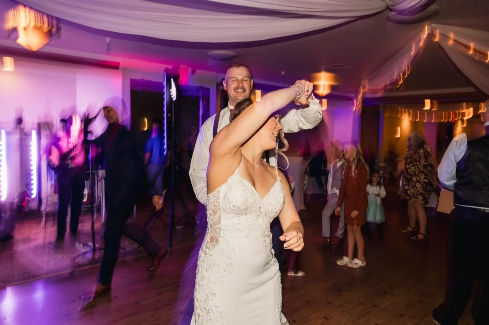 The groom spins the bride under his arm on a dance floor lit with purple uplights and draped ceiling fabric, guests dancing around them — Tim Larsen Photography, Brainerd Lakes MN