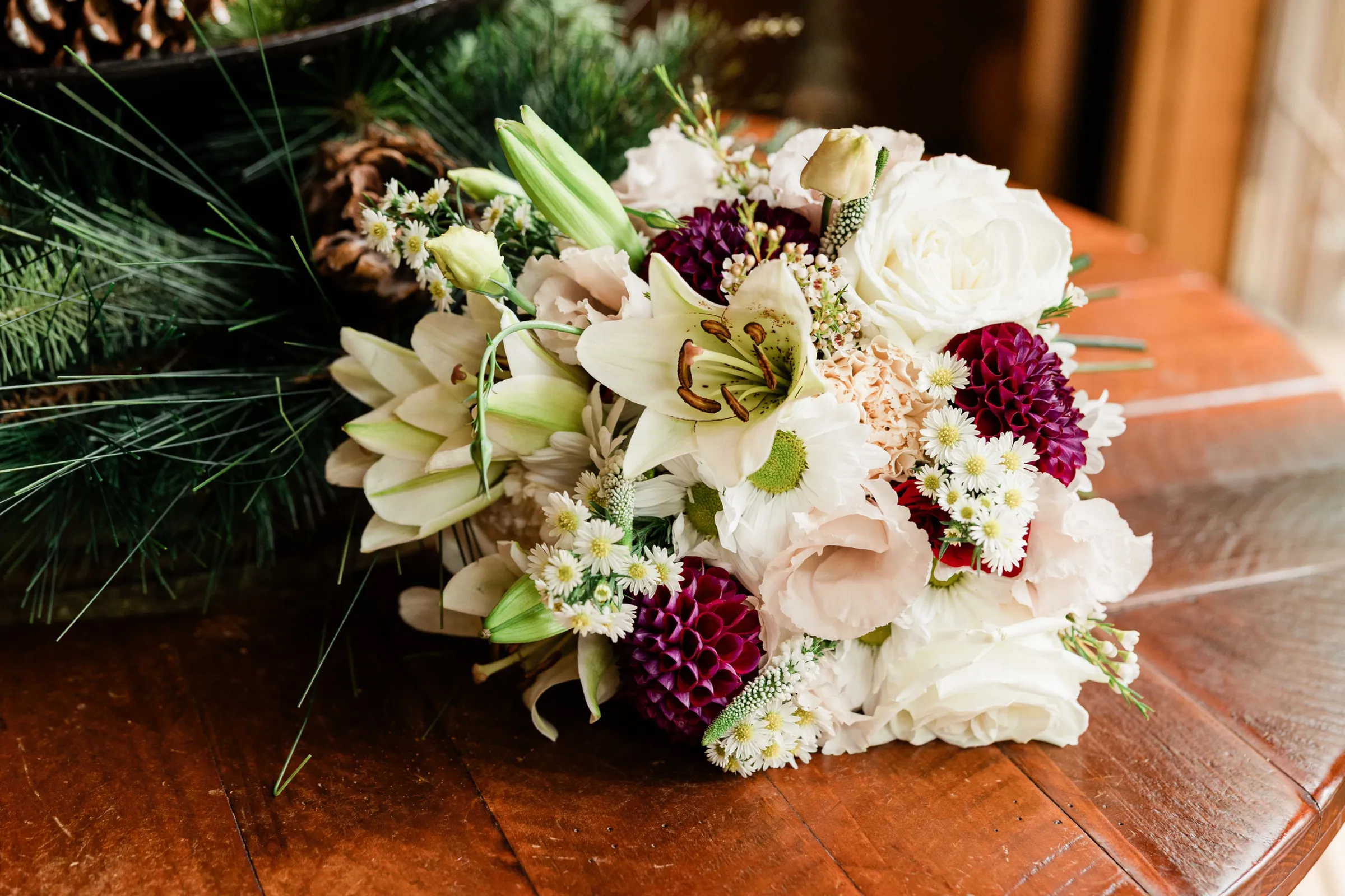 A bridal bouquet of burgundy dahlias, white lilies, roses, daisies and blush lisianthus lies on a round wooden table beside pine boughs and pinecones. — Tim Larsen Photography, Brainerd Lakes MN