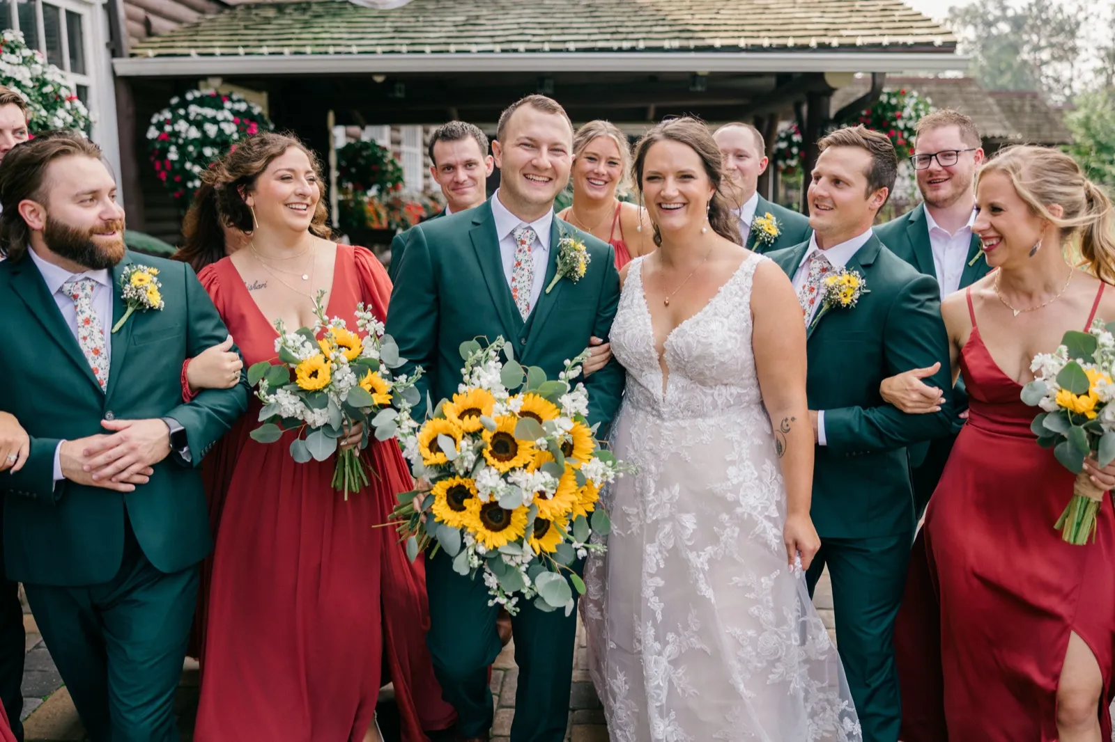 Chelsea and Eric walk forward arm-in-arm with their bridal party in green suits and red dresses, holding sunflower bouquets, in front of a flower-lined lodge porch — Tim Larsen Photography, Brainerd Lakes MN