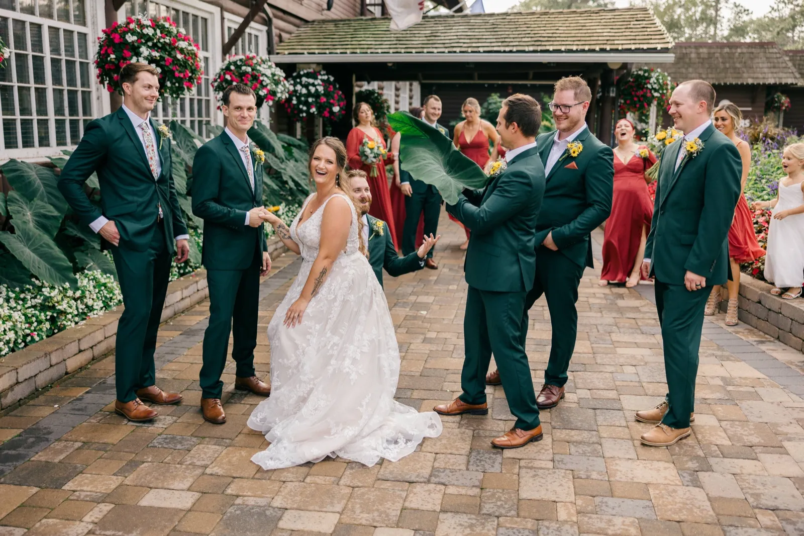 Chelsea laughs in the center of her wedding party as a groomsman waves a large tropical leaf beside her on a brick walkway outside Grand View Lodge — Tim Larsen Photography, Brainerd Lakes MN