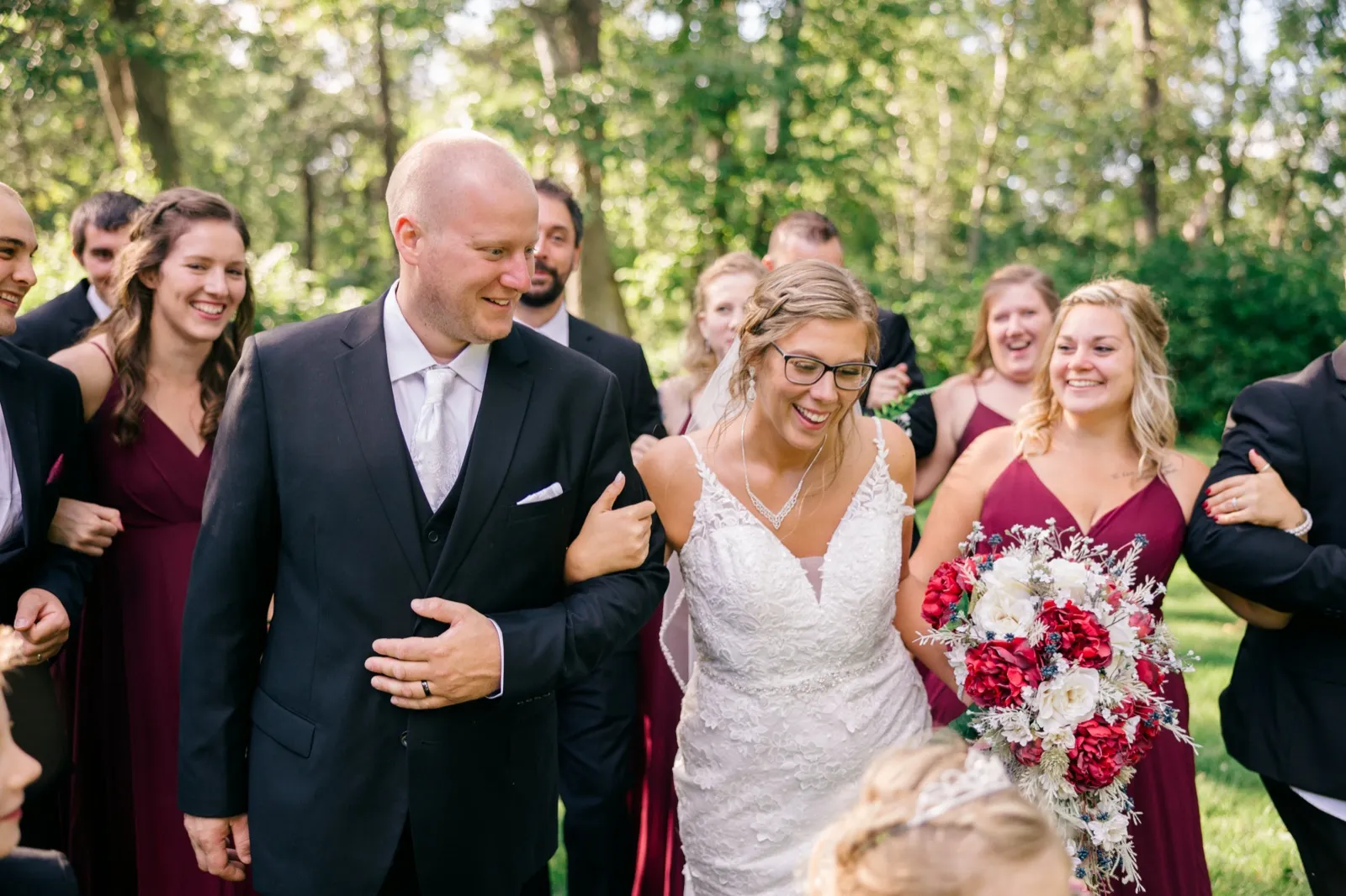 Kim laughs while holding a deep red and white bouquet as Jesse and the bridal party walk alongside through a sunlit wooded clearing — Tim Larsen Photography, Brainerd Lakes MN