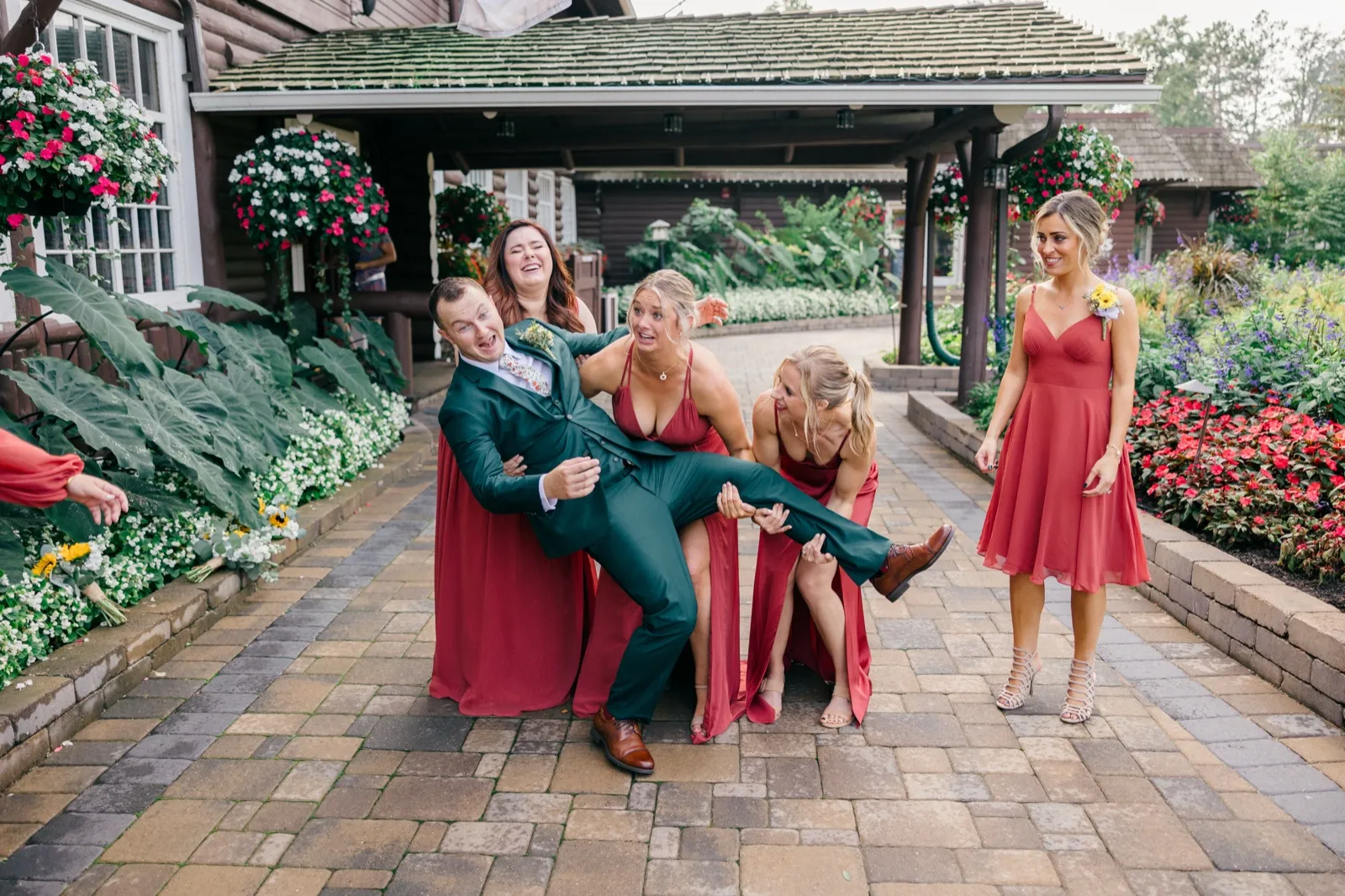 The bridal party lifts Eric horizontally across their arms while laughing on a paver walkway outside Grand View Lodge, surrounded by hanging flower baskets and gardens — Tim Larsen Photography, Brainerd Lakes MN