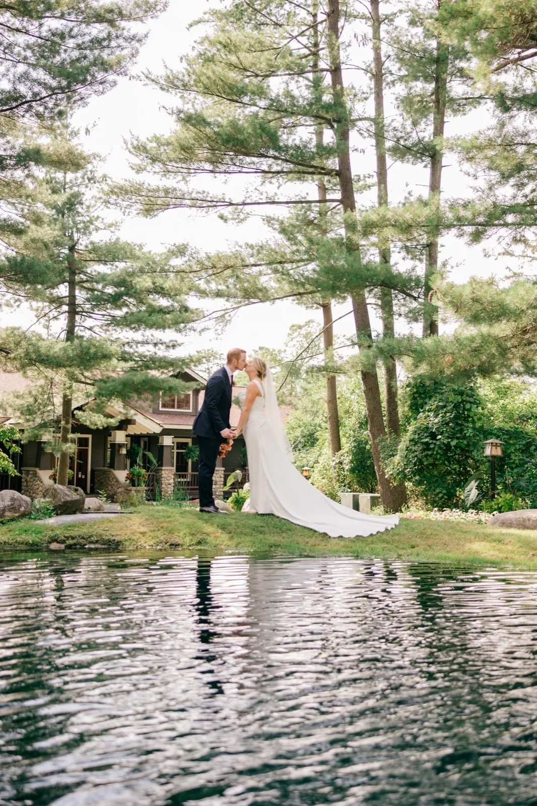 Monika and Mats stand together on a lawn beside a pond, the bride's long train trailing across the grass beneath towering pine trees — Tim Larsen Photography, Brainerd Lakes MN