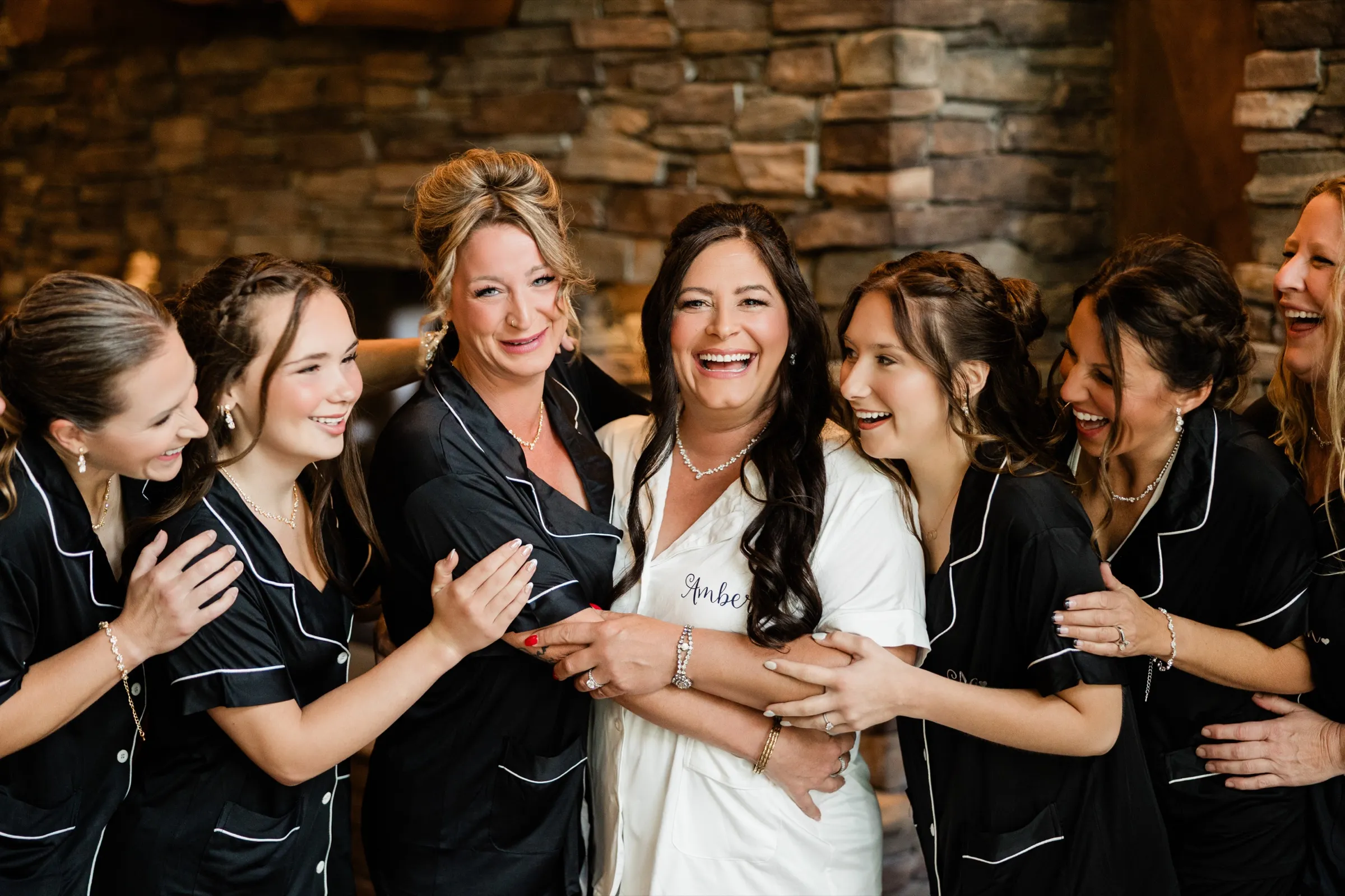 The bride throws her head back laughing while the women around her in black pajamas lean in, laughing and holding her arms, in front of a stone wall. — Tim Larsen Photography, Brainerd Lakes MN