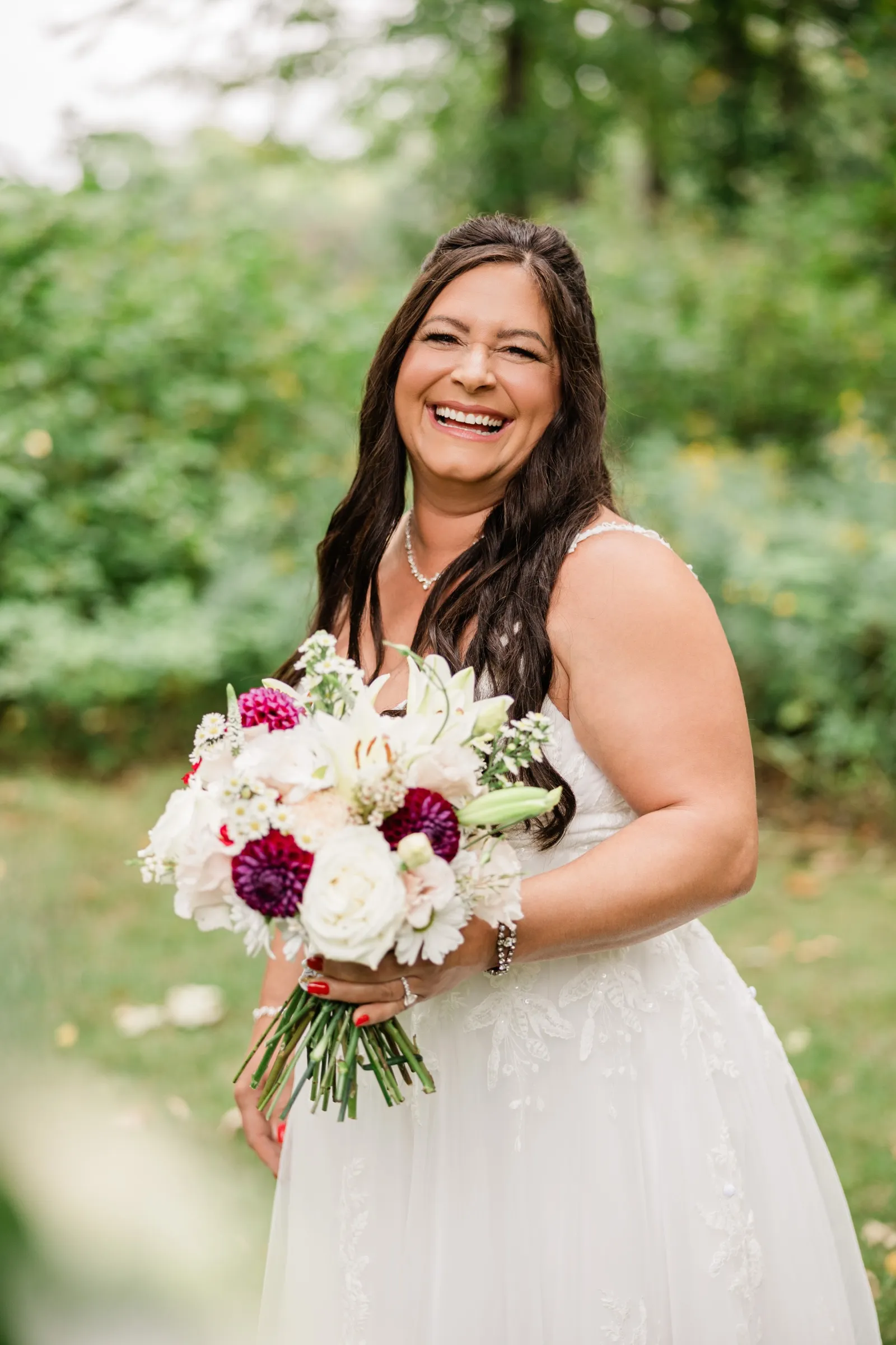 The bride laughs openly while holding her bouquet of burgundy dahlias, white lilies and roses against a soft green background. — Tim Larsen Photography, Brainerd Lakes MN