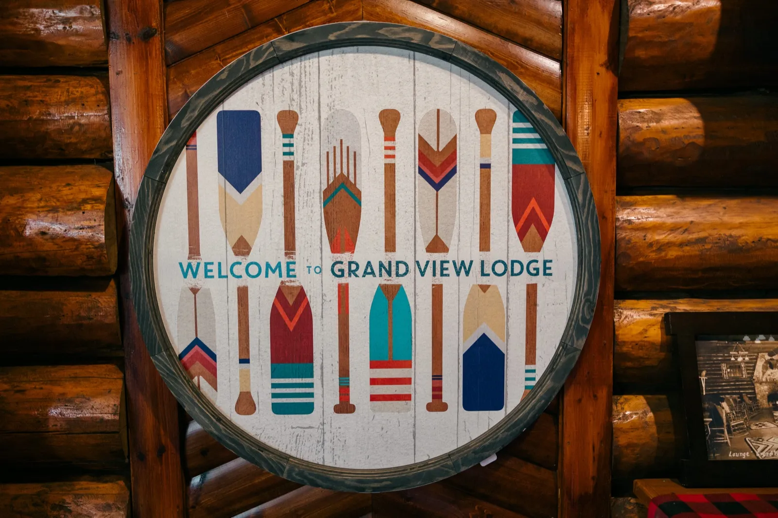A round wooden sign painted with colorful canoe paddles reads 'Welcome to Grand View Lodge,' mounted on a log cabin wall — Tim Larsen Photography, Brainerd Lakes MN