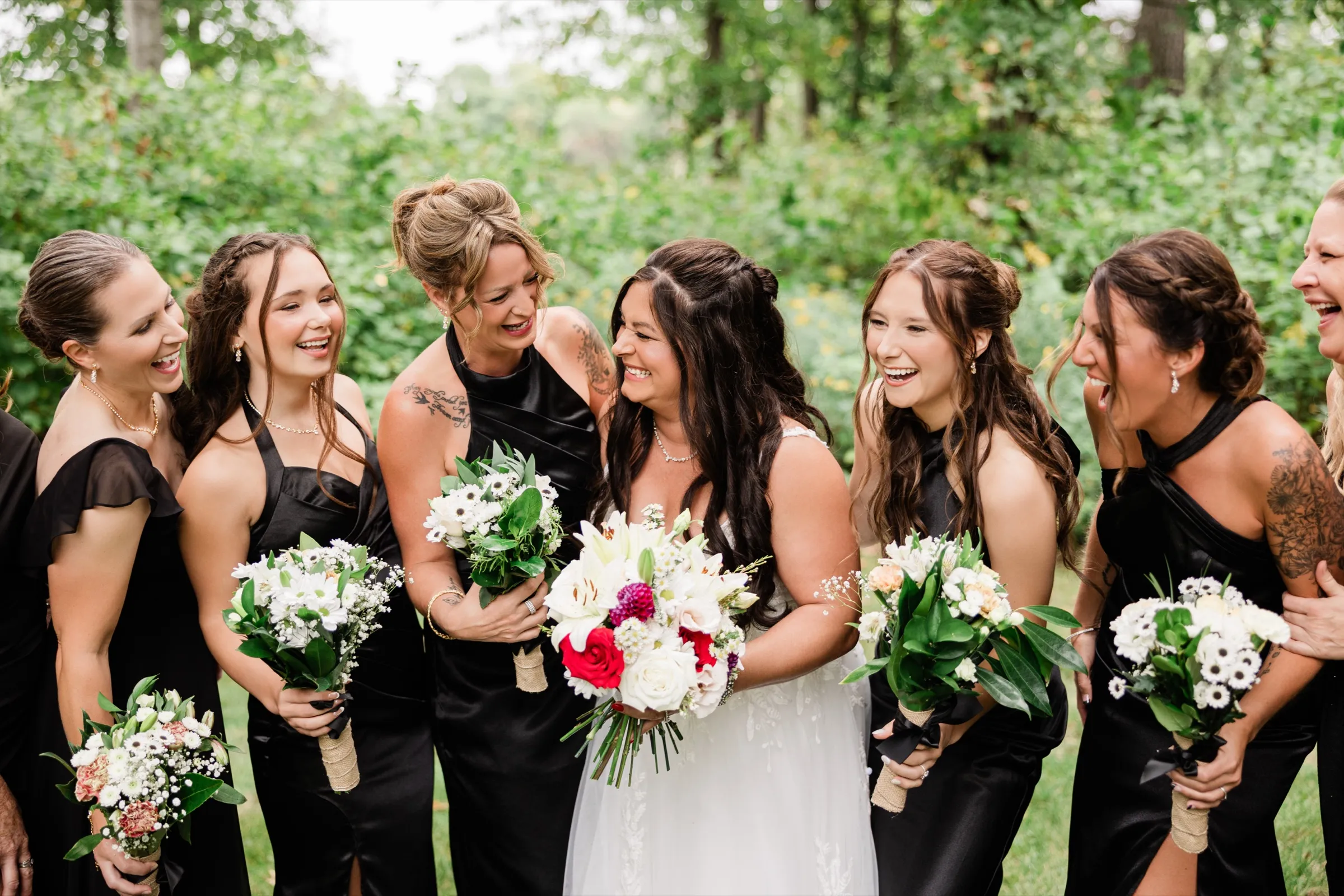 The bride and bridesmaids in black satin dresses laugh together on the lawn, holding white bouquets while the bride holds hers of red roses and white lilies. — Tim Larsen Photography, Brainerd Lakes MN