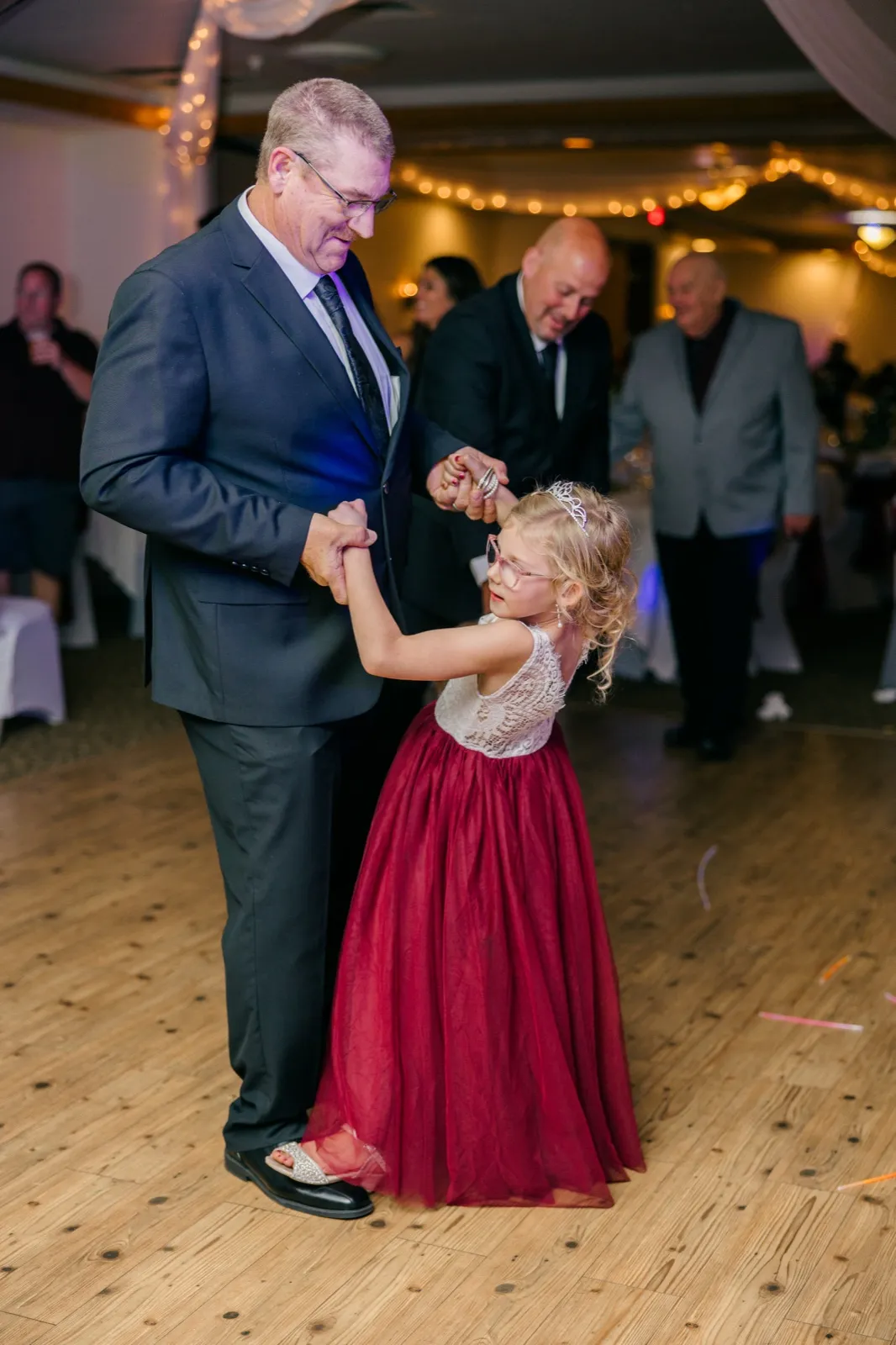A man in a gray suit twirls a young flower girl in a burgundy tulle gown and tiara on the reception dance floor — Tim Larsen Photography, Brainerd Lakes MN