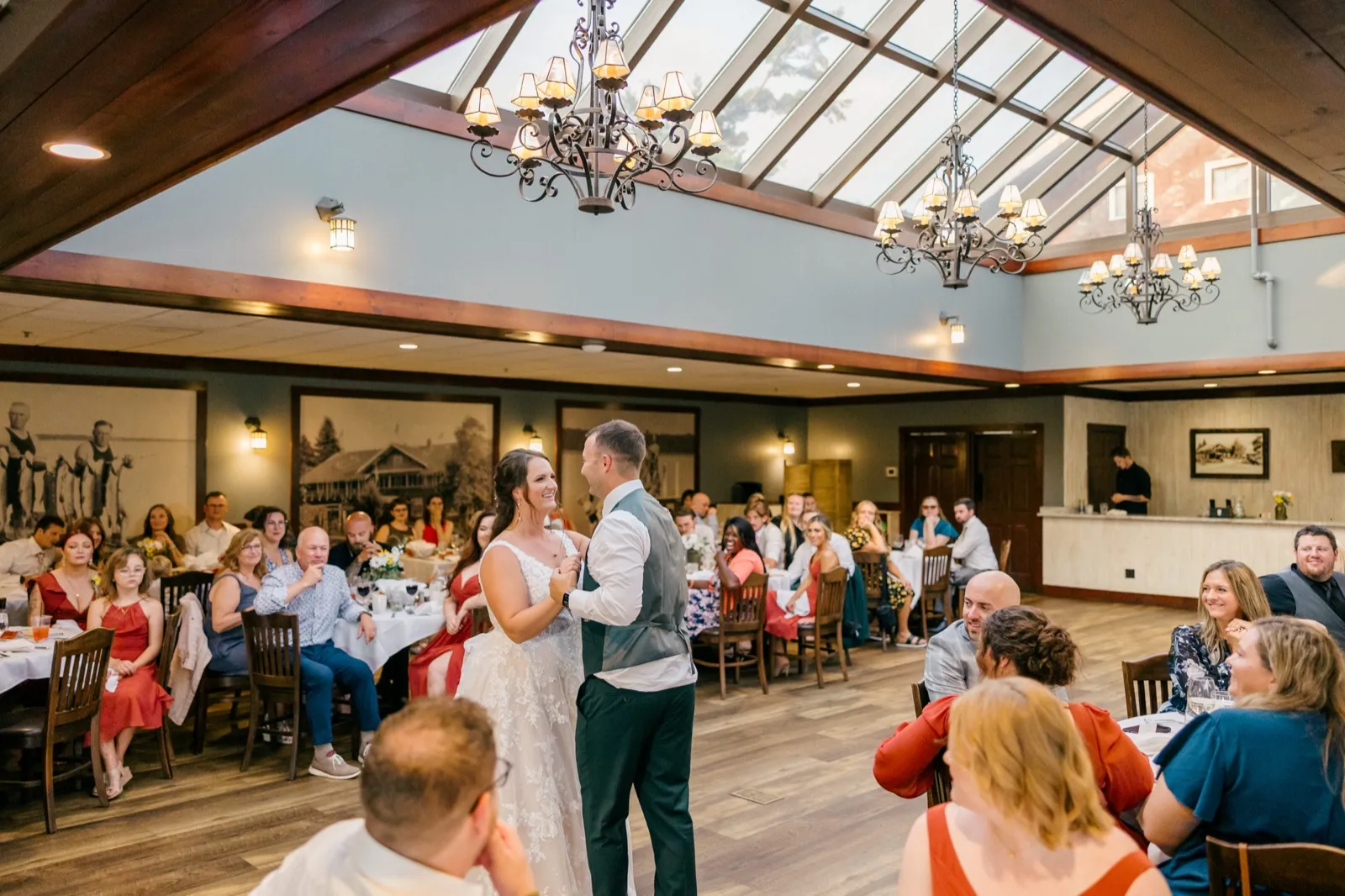 Chelsea and Eric share their first dance in a skylit ballroom with chandeliers overhead and seated guests watching from tables on either side — Tim Larsen Photography, Brainerd Lakes MN