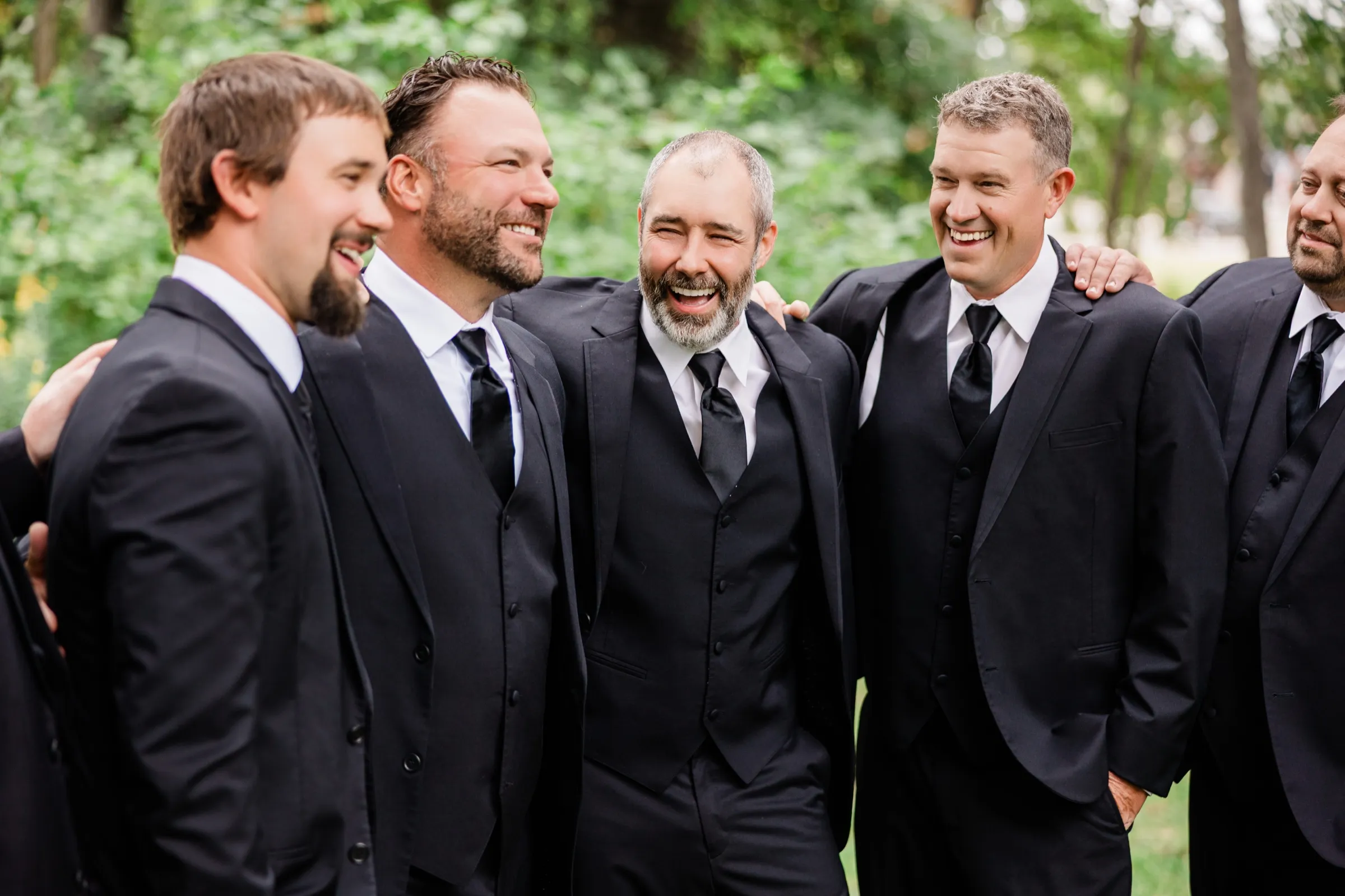 The groom laughs with his eyes squeezed shut as groomsmen in black suits drape their arms over his shoulders and grin beside him. — Tim Larsen Photography, Brainerd Lakes MN